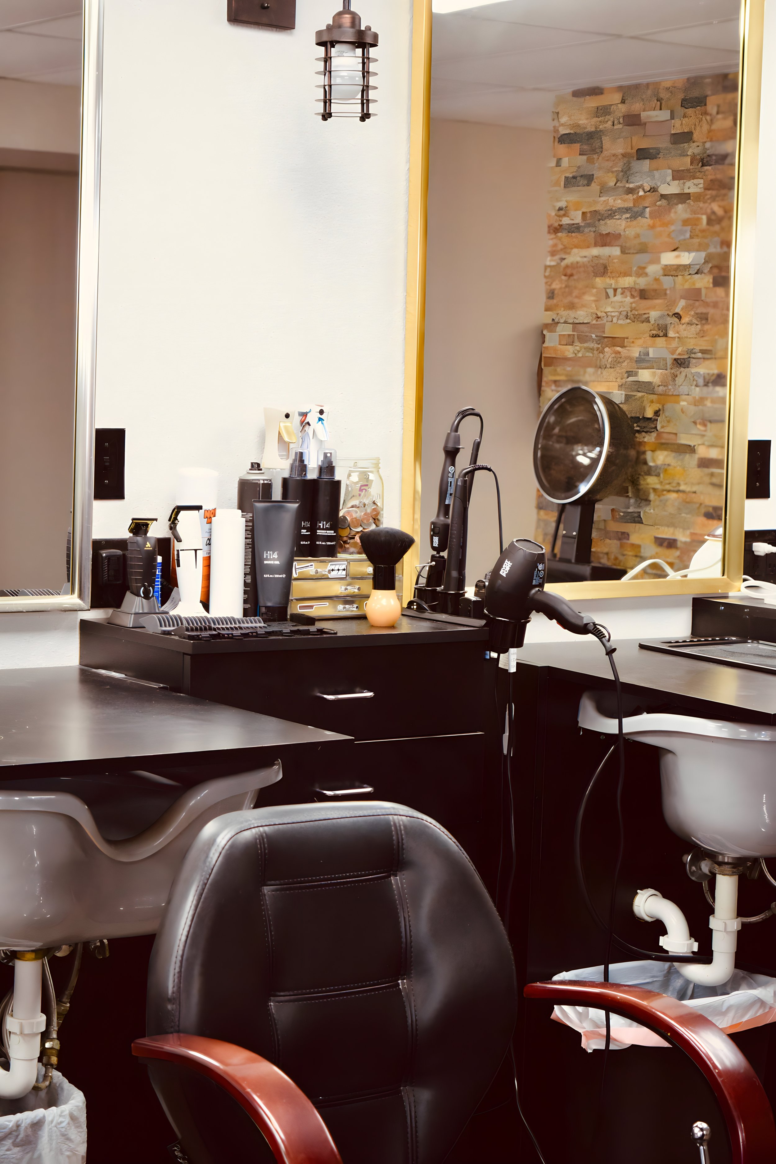 A hair salon station with black drawers, a mirror, and various hair care products, including sprays, creams, and a styling brush. There are chairs and sinks for hair washing, and hair dryers are attached to the counter.