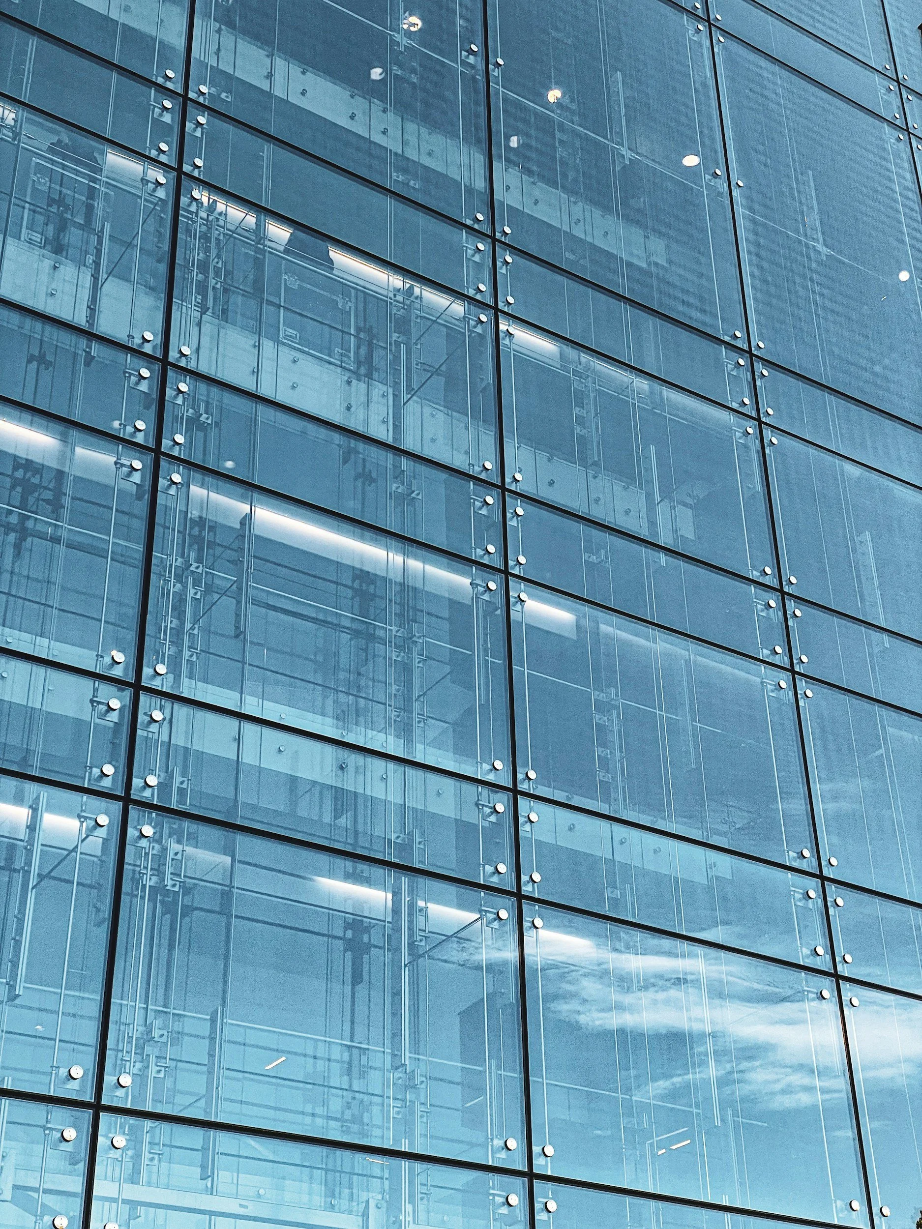 Close-up of a glass building facade with reflections of the sky and clouds, and visible interior structures behind the glass.