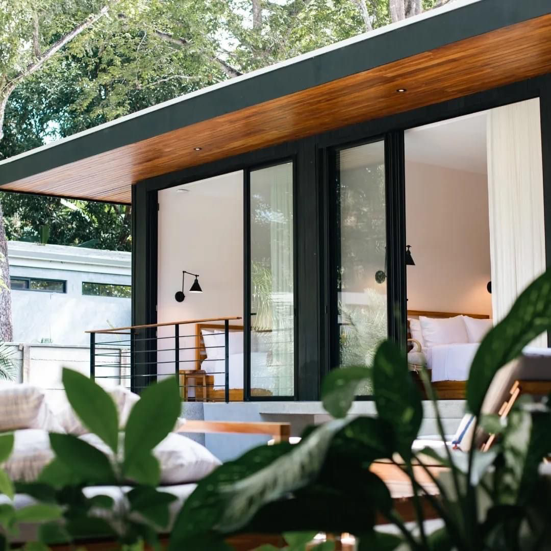 Modern house with large glass sliding doors, wooden accents, and a cozy interior featuring a bed with white linens, surrounded by lush greenery.