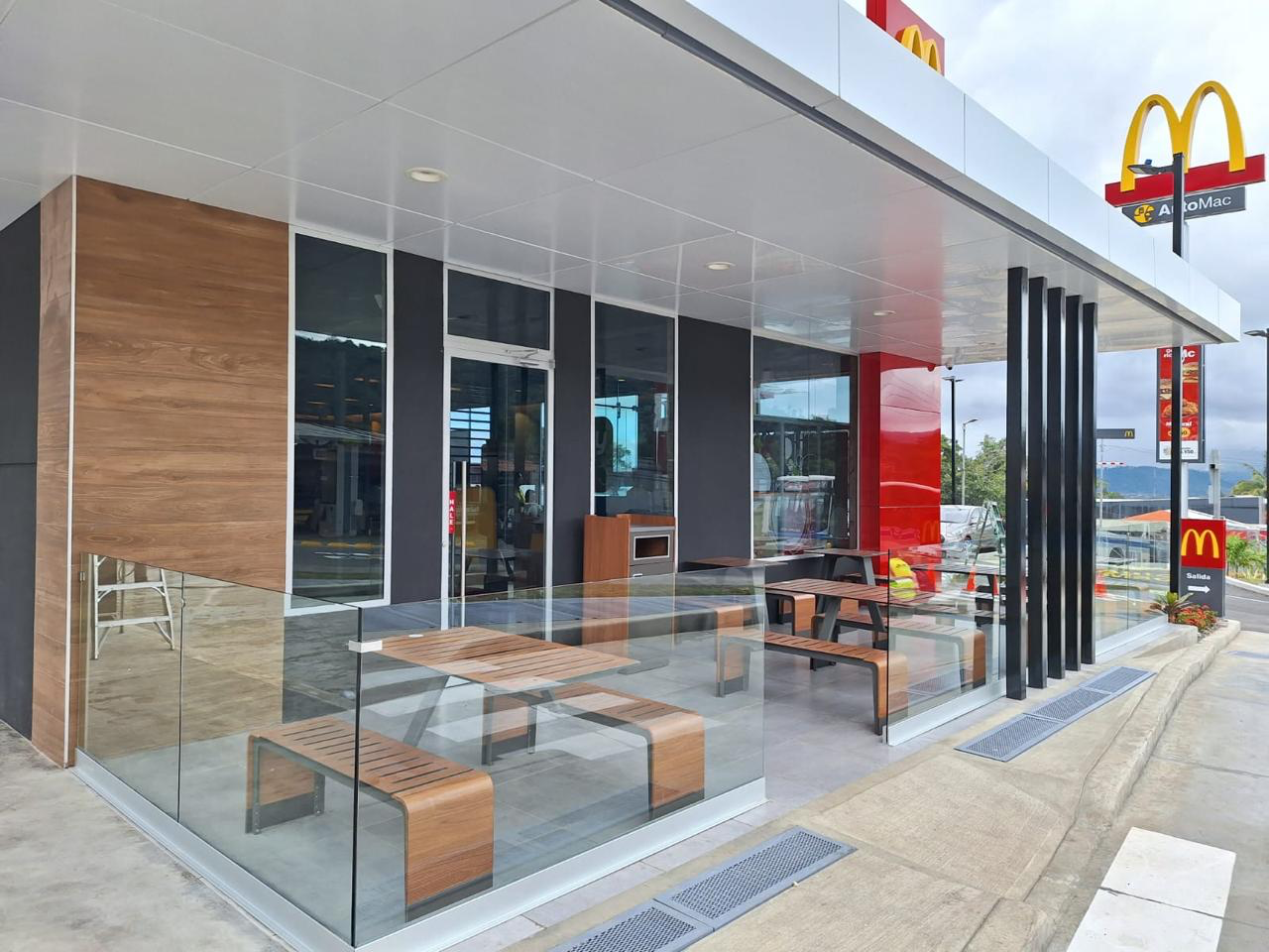 Exterior view of a McDonald's restaurant with a glass-walled outdoor seating area, wooden benches, a red accent wall, and McDonald's signage.