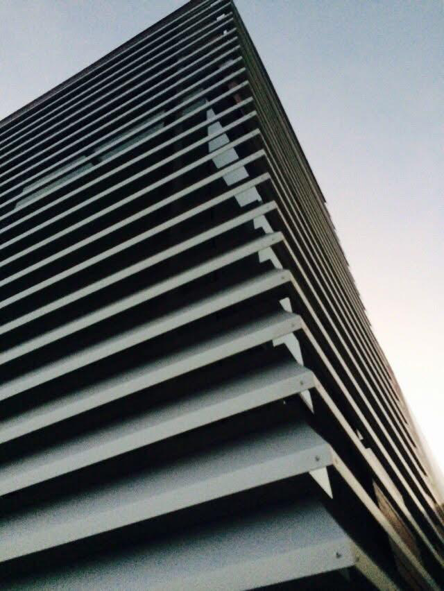 Looking up at a modern high-rise building with a pattern of horizontal metal louvers or slats, against a clear sky.