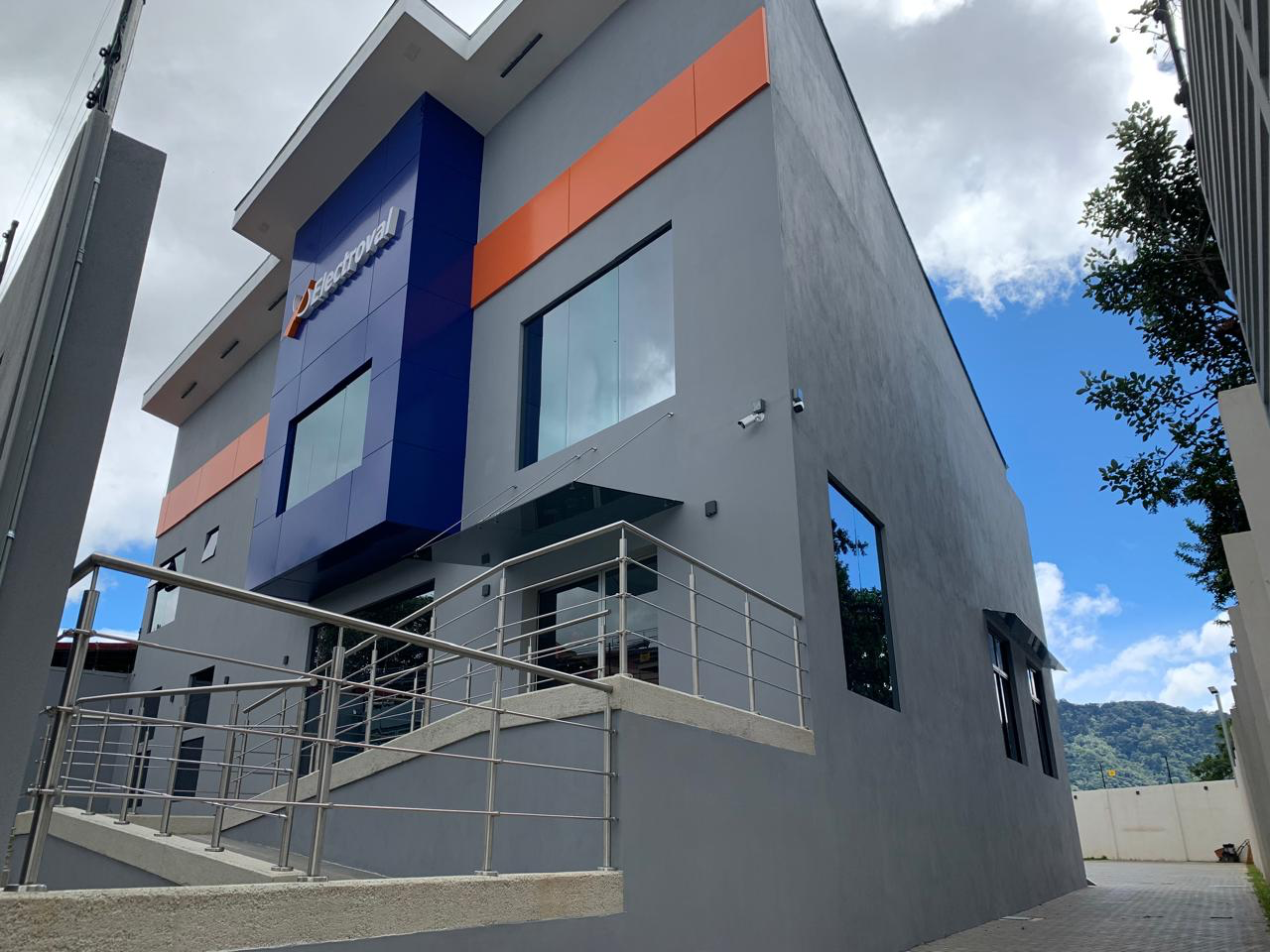 Modern commercial building with blue, orange, and gray exterior, Large glass windows, Railing on stairs, Company logo on top left corner, Mountain in background.
