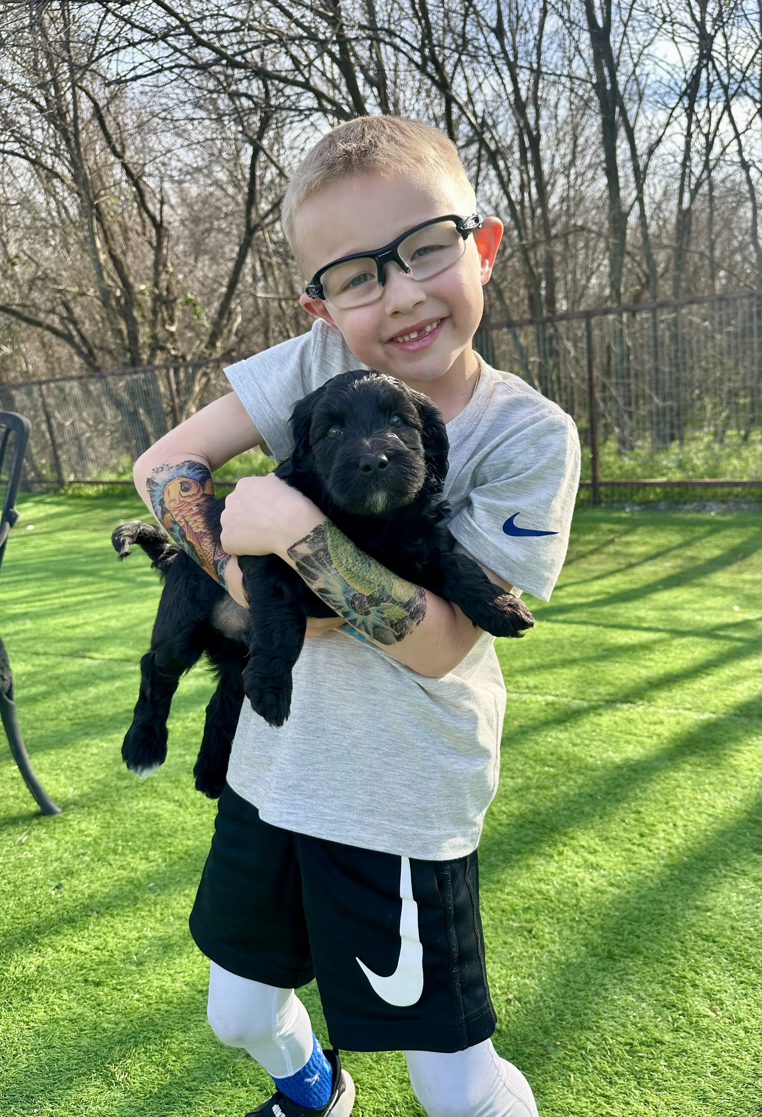 A young boy wearing glasses, a gray Nike t-shirt, black Nike shorts, and white compression sleeves, holding a black puppy outdoors on a sunny day with green grass and trees in the background.