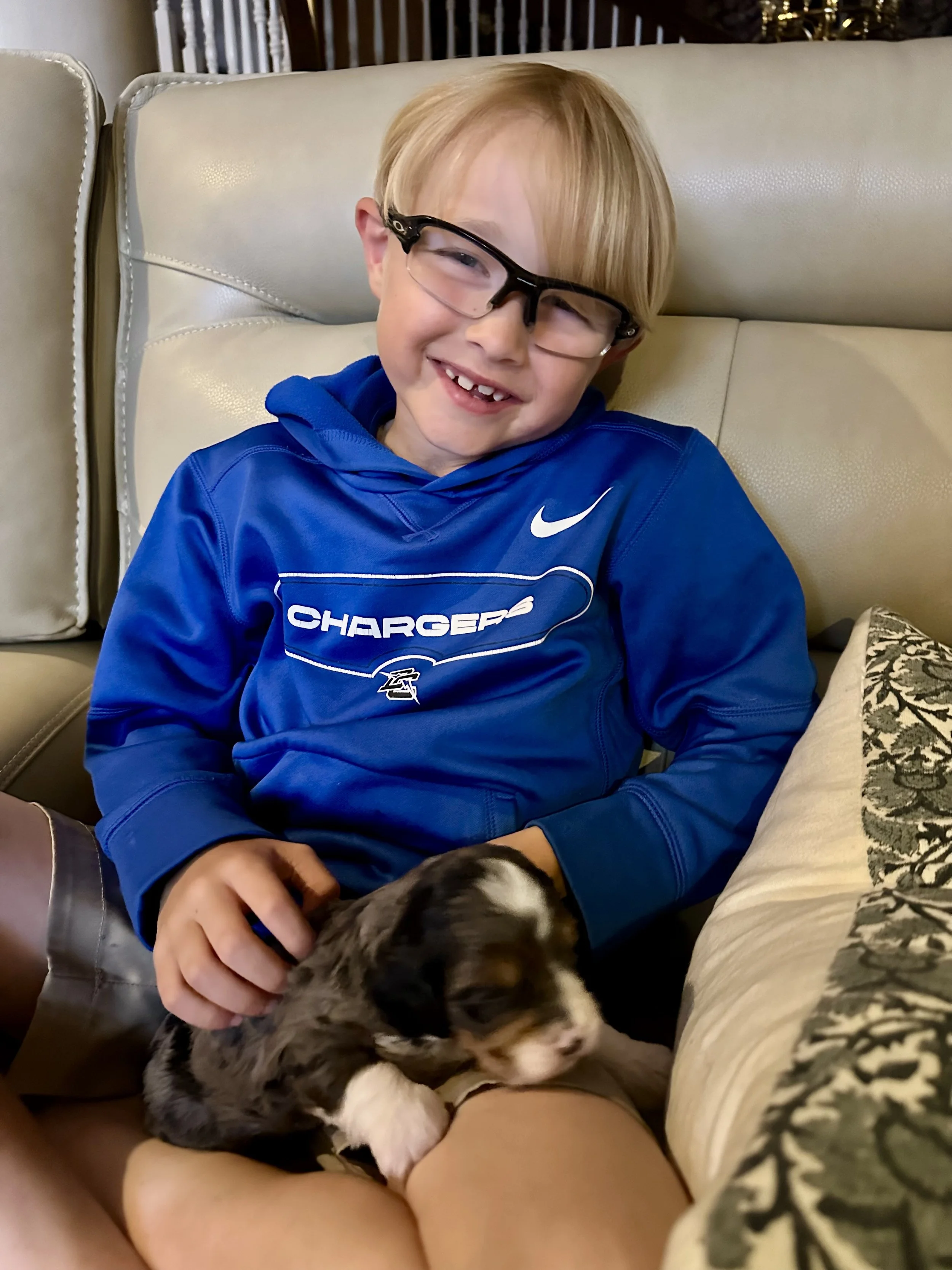 A young boy with glasses and a missing tooth, smiling and sitting on a couch, holding and gently petting a small puppy.