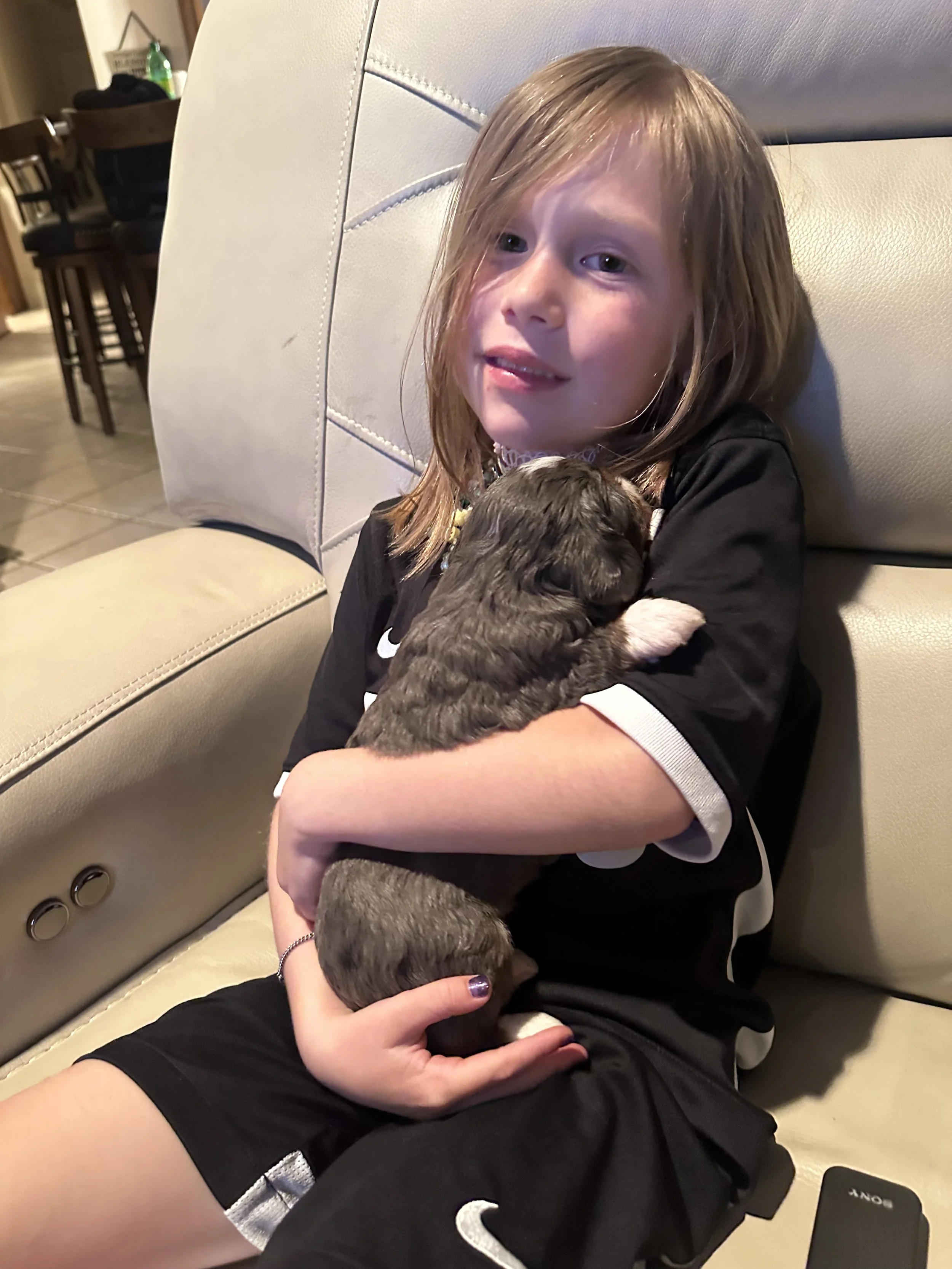 A young girl with shoulder-length light brown hair, wearing a black sports jersey, sitting on a beige leather couch, holding a small, dark-colored puppy.