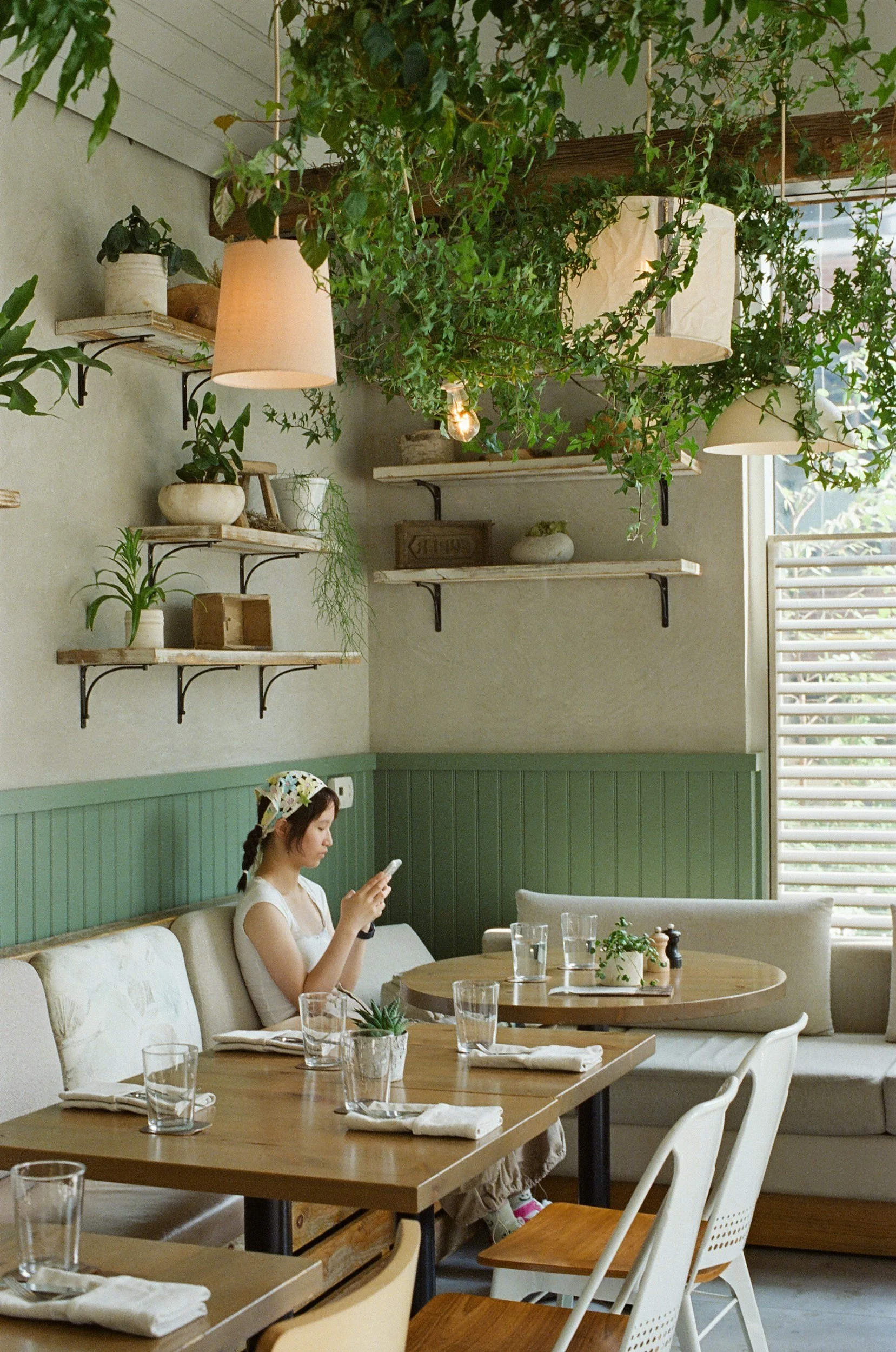 A woman sitting at a dining table in a cozy, green-themed restaurant, shelves of faux potted plants and hanging greenery overhead.