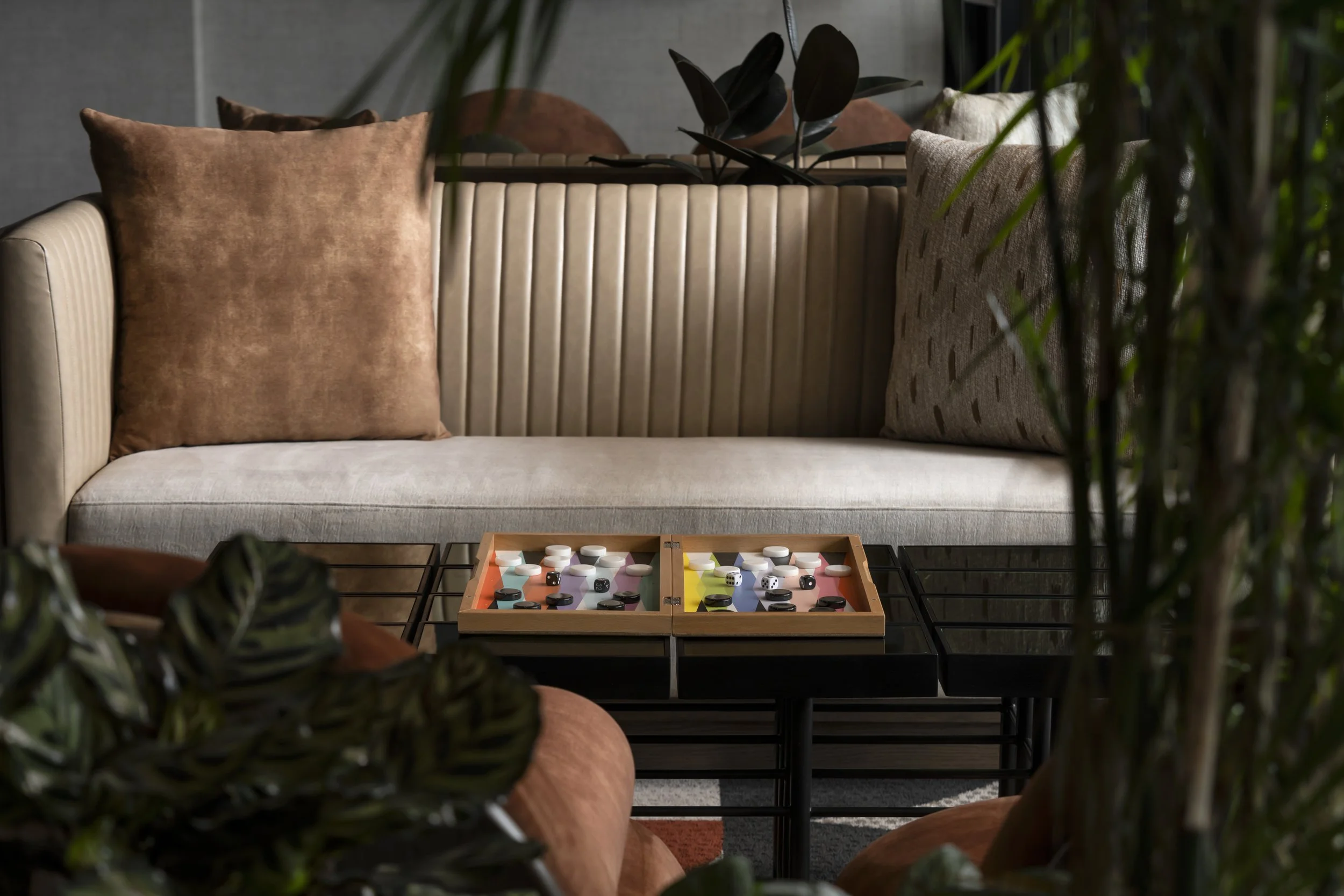 A cozy living room with beige upholstered sofa, adorned with textured pillows in shades of brown and beige, situated behind a black coffee table. The table holds a colorful backgammon game with black and white dice and chips, surrounded by indoor plants partially framing the scene.