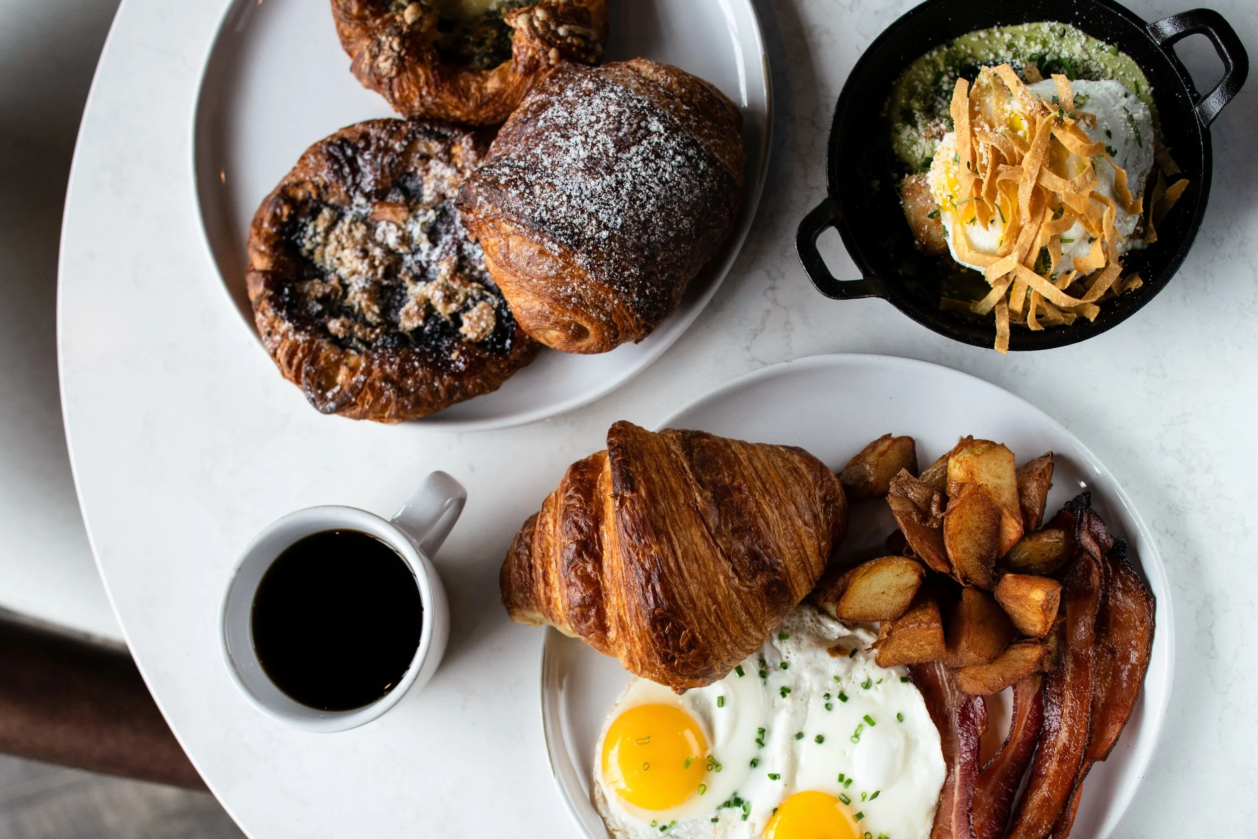 A breakfast table with a plate of croissants, a cup of coffee, and a breakfast platter with eggs, bacon, roasted potatoes, and a small skillet with a salad.