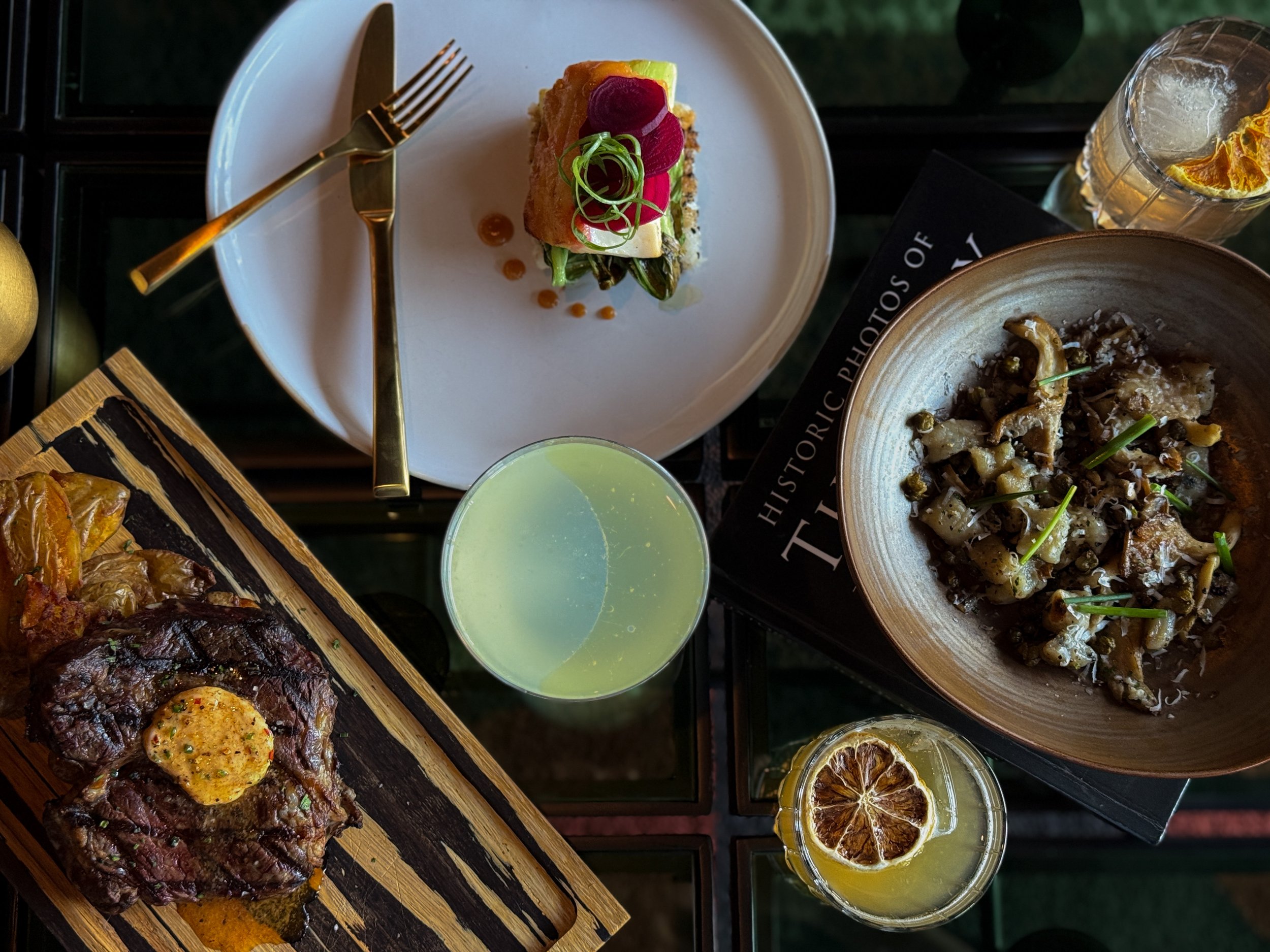 A variety of dishes on a black table, including a grilled steak with a pat of herb butter, a colorful layered vegetable tower, a bowl of pasta with mushrooms, and a glass of lemon water with dried orange slices.