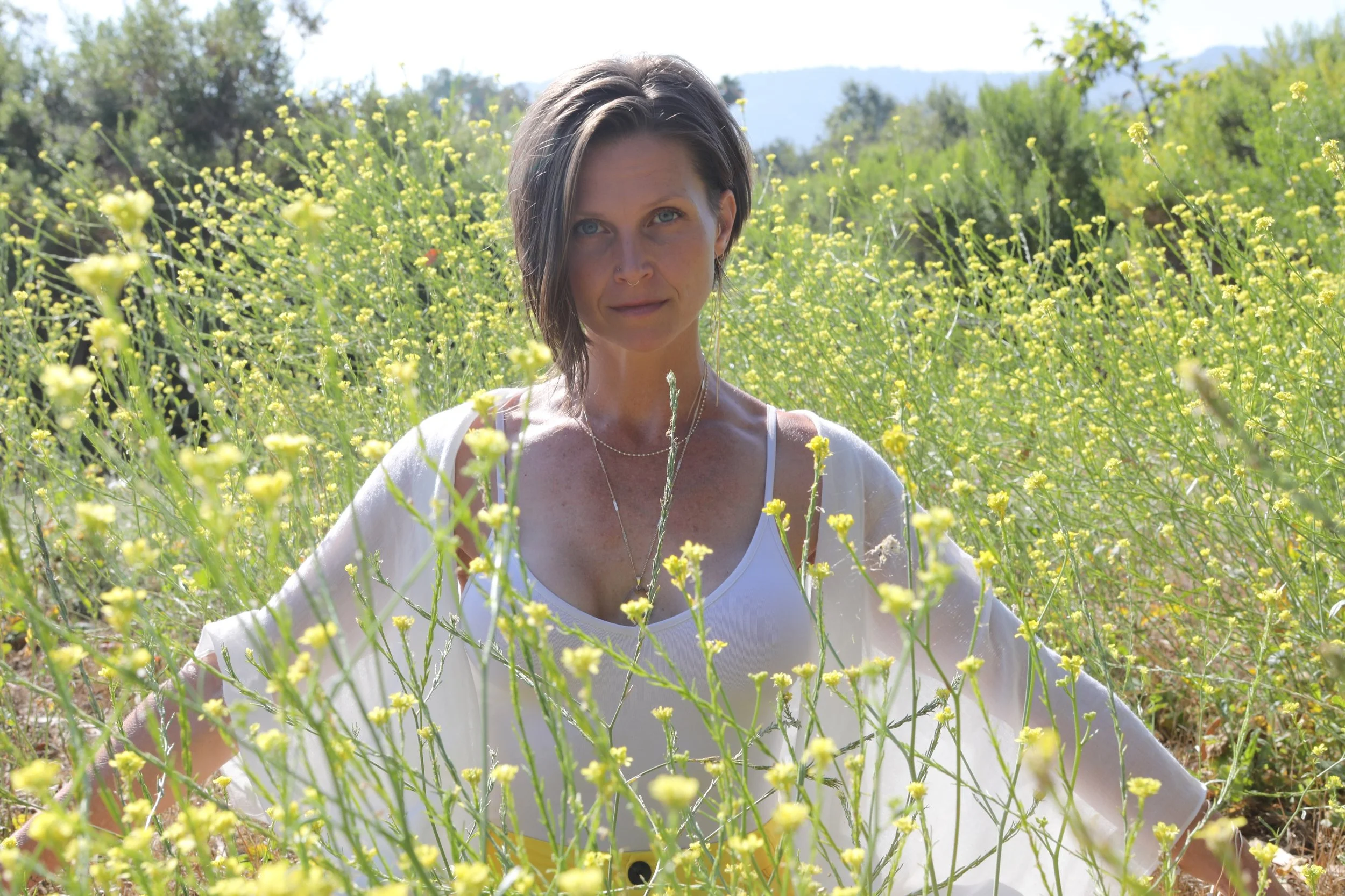 A woman with short brown hair and blue eyes standing in a field of yellow flowers on a sunny day, wearing a white tank top and a sheer white cover-up.