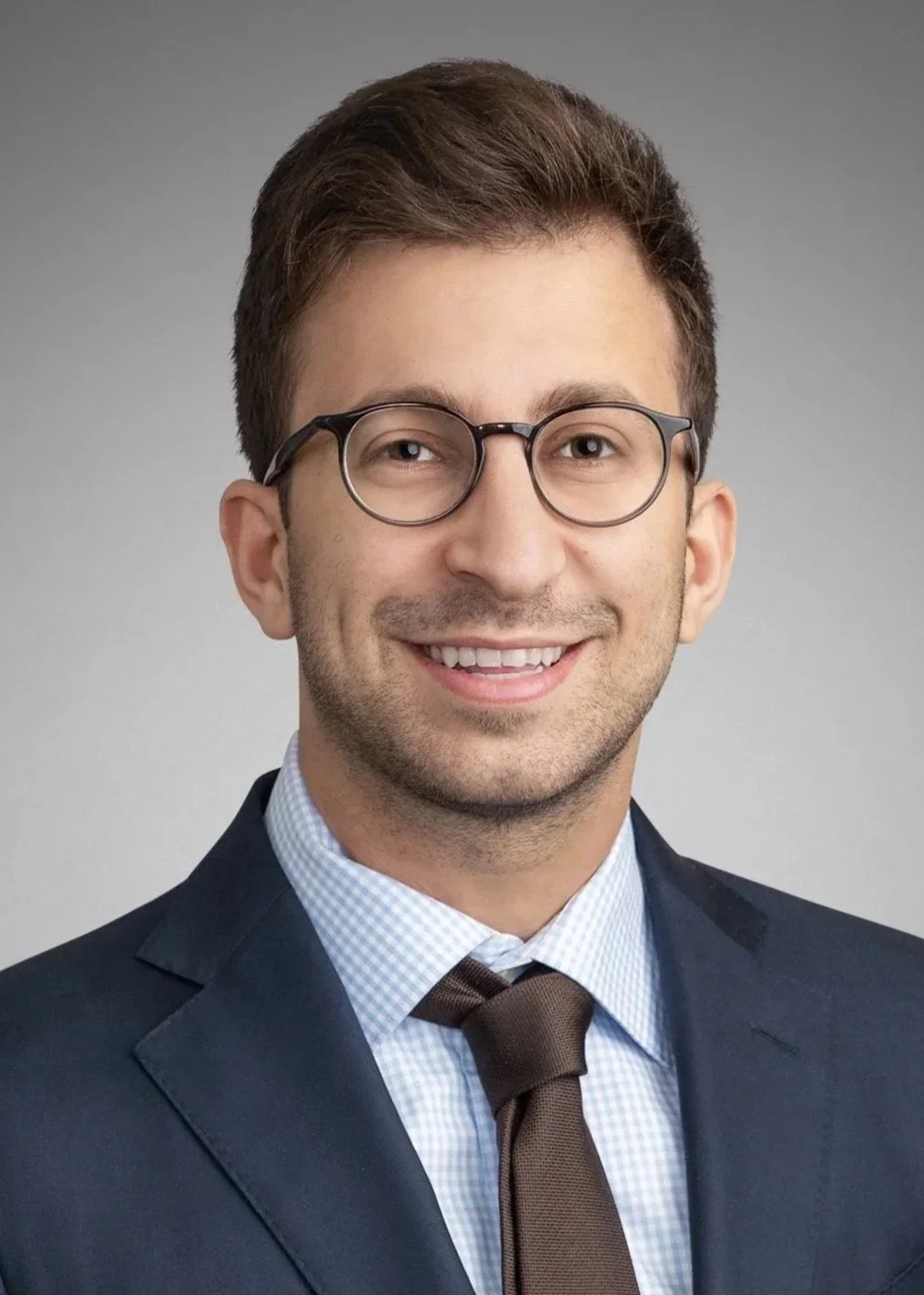 A professional headshot of a young man with brown hair, glasses, wearing a navy blue suit, light blue checked shirt, and a brown tie, smiling against a plain gray background.