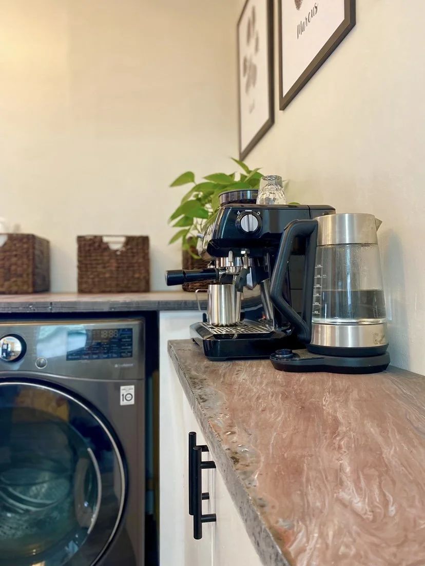 Close-up of a custom coffee bar featuring a solid wood countertop with espresso machine and electric kettle, integrated into built-in cabinetry alongside a laundry area, highlighting functional finish carpentry and workspace design.