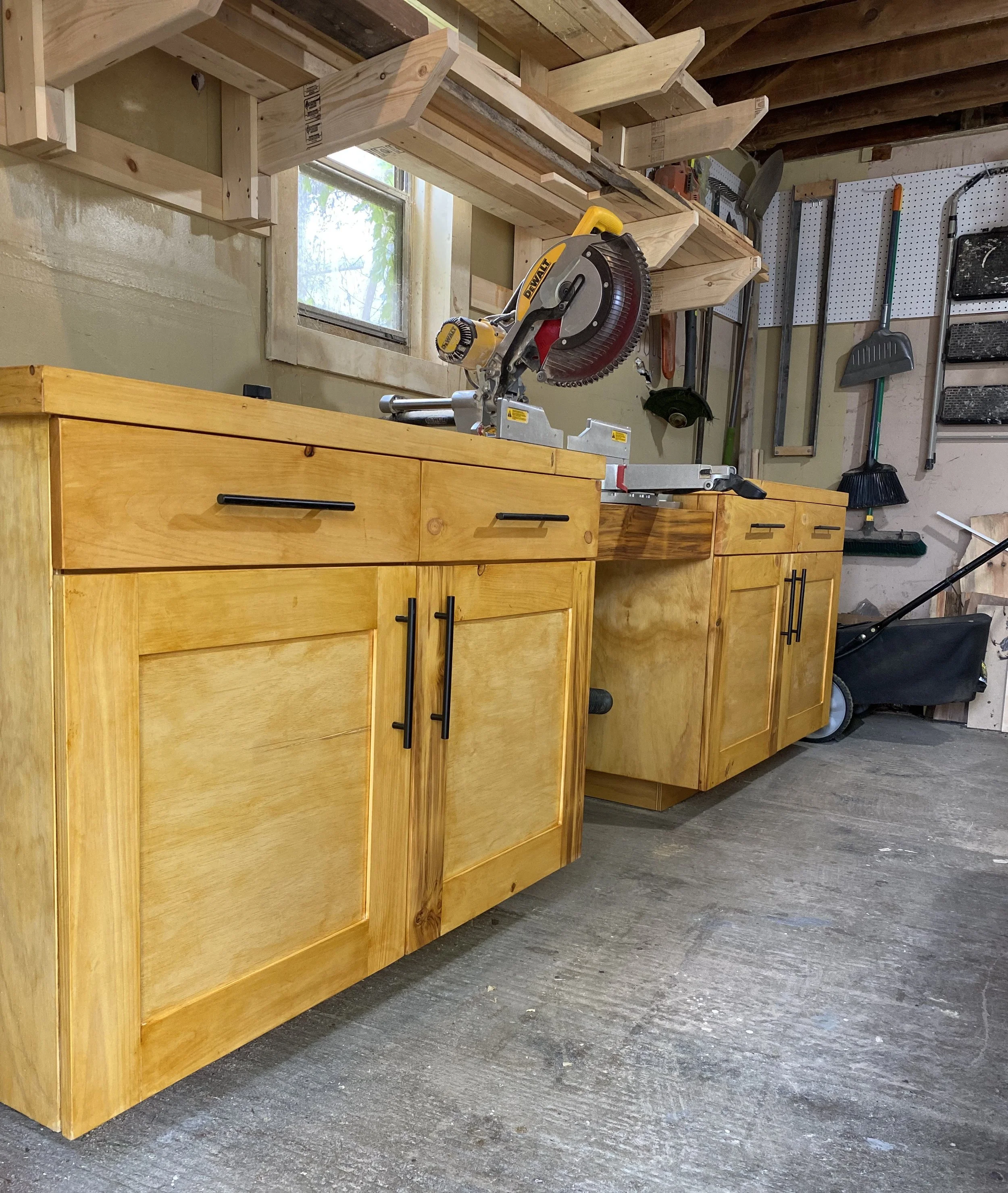 Custom wood base cabinets with shaker-style doors and black hardware shown in a Beverwyck Designs woodworking shop beneath a built-in miter saw workstation.