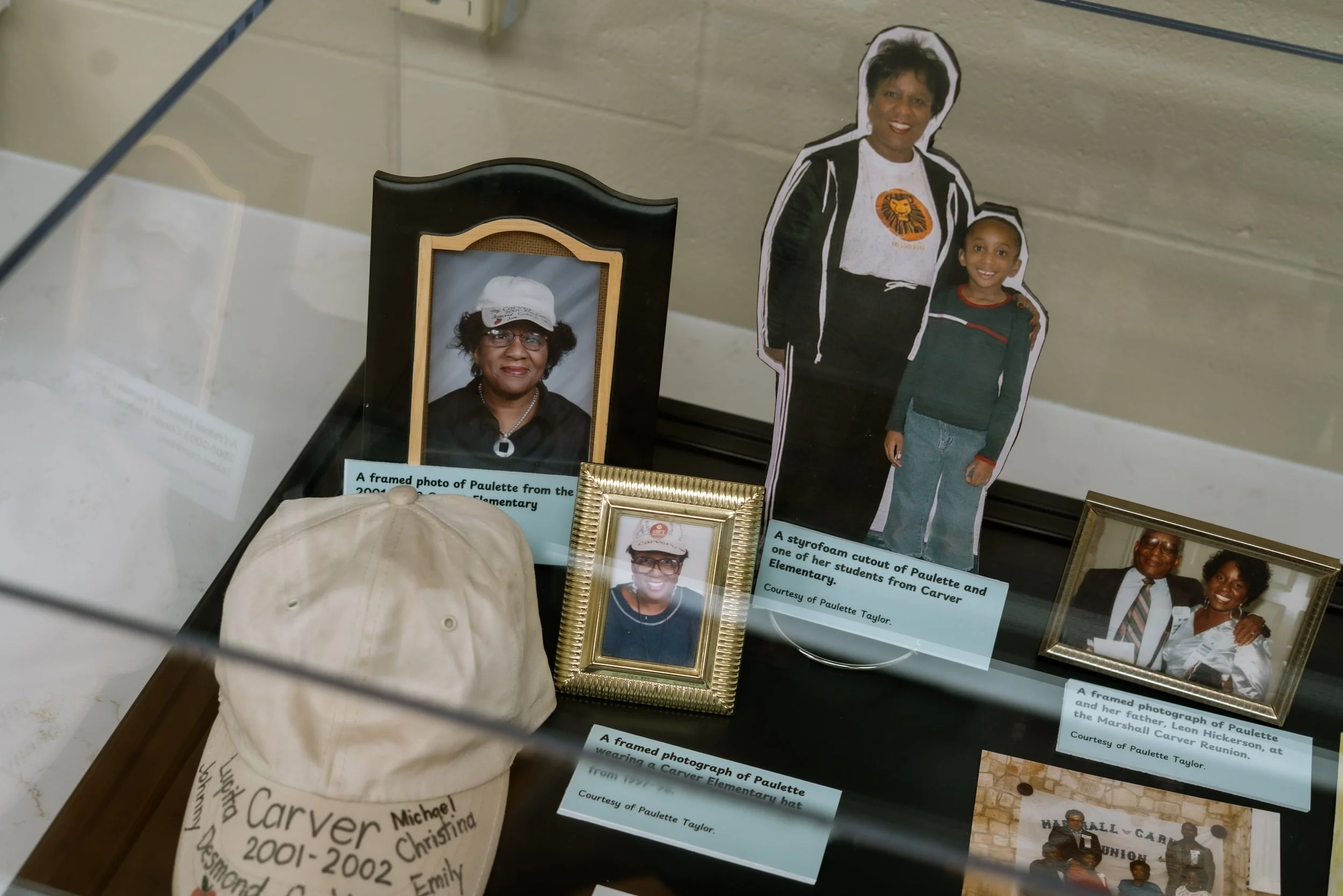 Display case with framed photographs and memorabilia of Paulette from elementary school, including a cutout of Paulette and a woman, possibly her mother, in a family reunion photo, along with descriptive labels and captions.