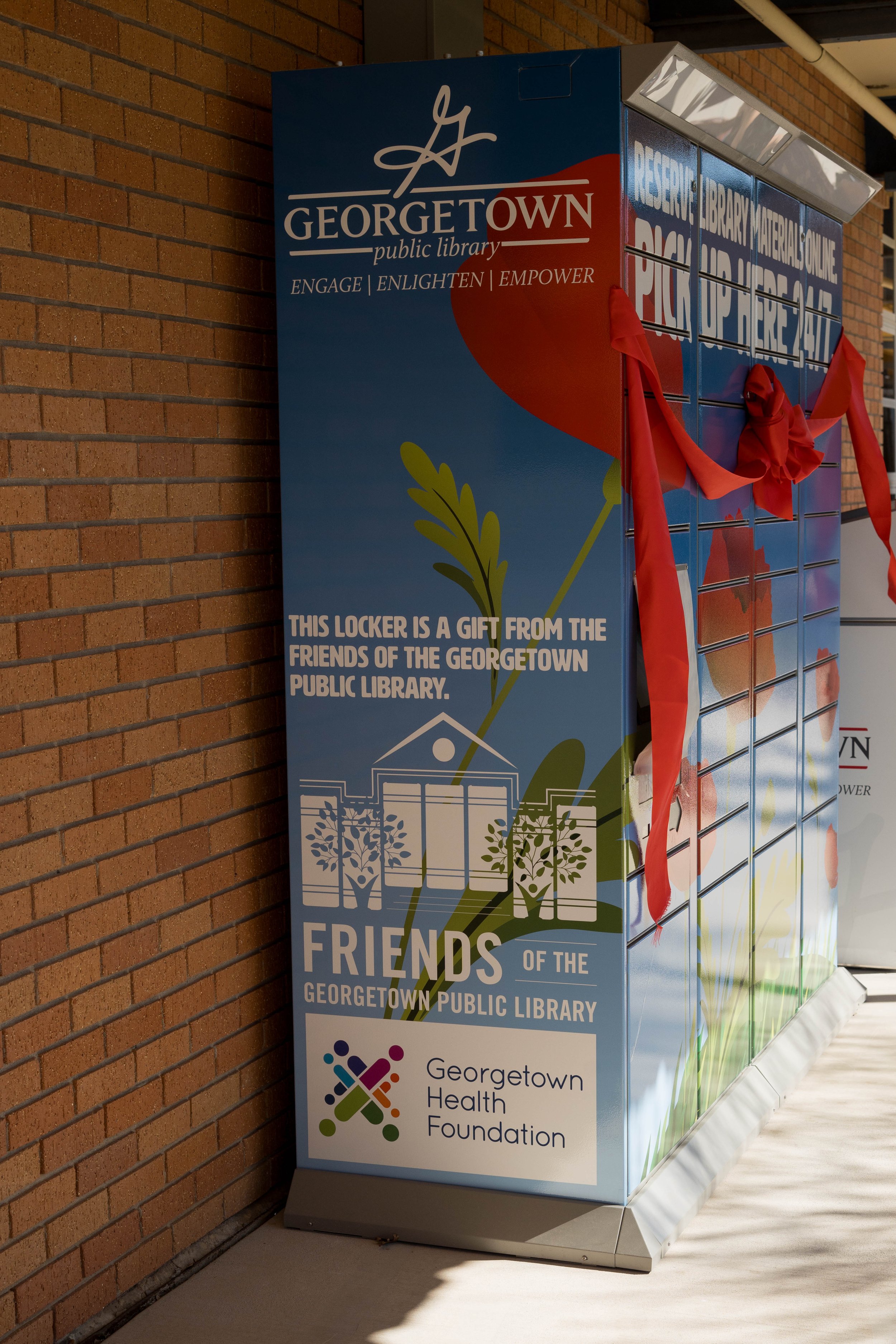 A colorful outdoor locker with multiple compartments decorated with red ribbons and flowers, placed next to a brick wall, sponsored by the Georgetown Public Library and Georgetown Health Foundation.