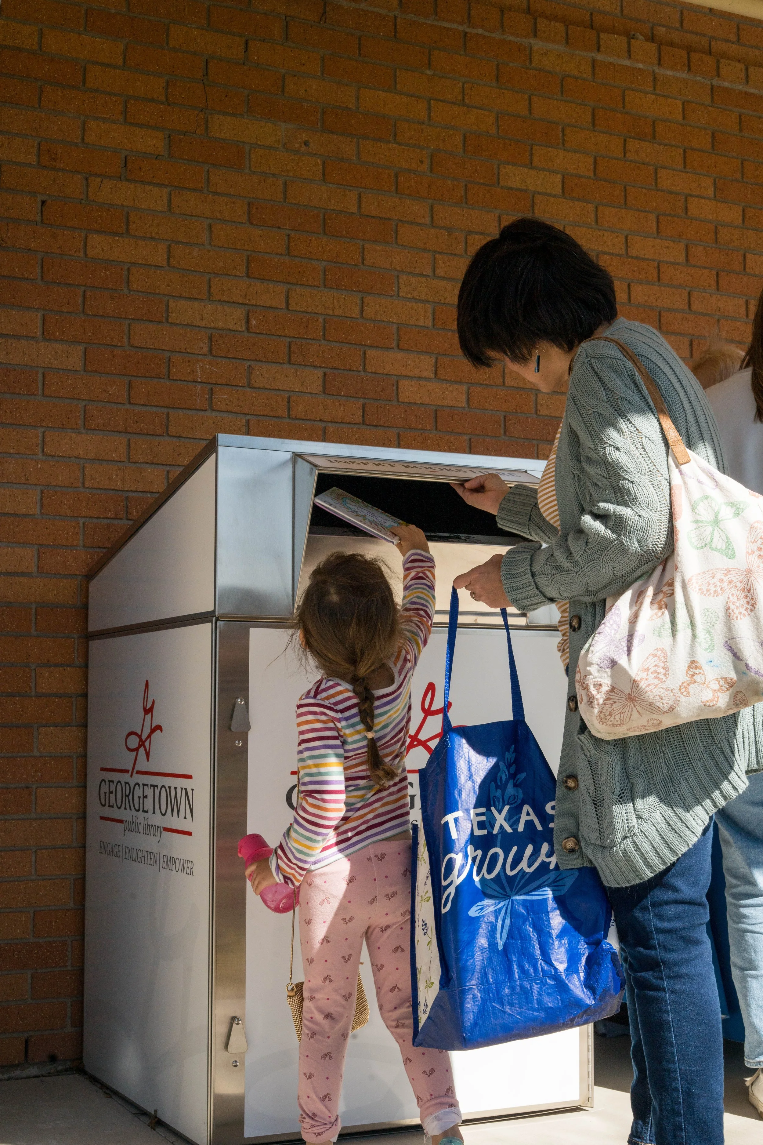 A woman and a child are returning books at a public library, Georgetown. The woman wears a gray cardigan and carries a blue shopping bag, while the girl wears pink pajamas with butterfly prints and holds a pink water bottle.