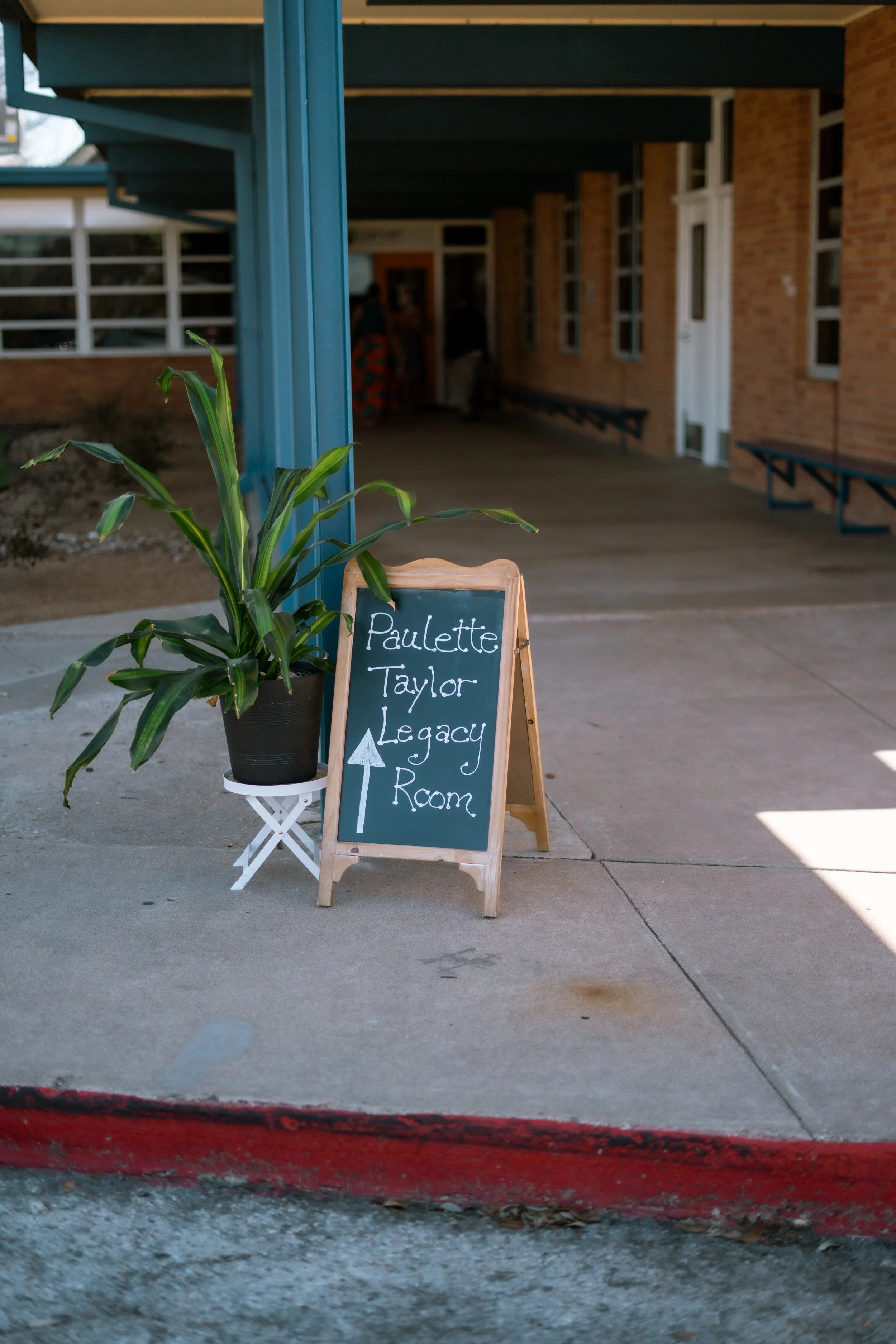 A potted plant with green leaves and long, narrow leaves stands next to a wooden sign on a sidewalk. The sign reads, 'Paulette Taylor Legacy Room' with an arrow pointing upward. In the background, a school building with brick walls and multiple windows is visible, along with a covered walkway and people inside.
