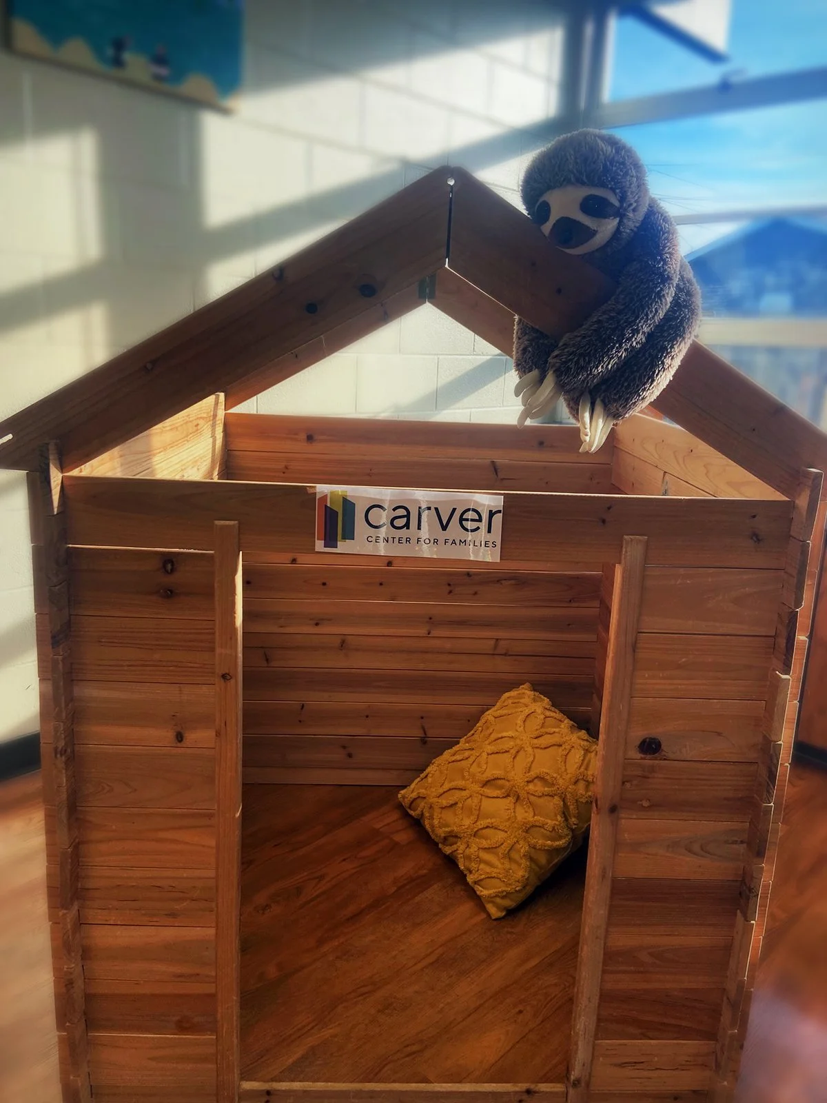 A wooden doghouse with a sloped roof, inside is a yellow pillow with a textured pattern. A plush sloth toy is draped over the roof, and there is a logo for Carver Center for Families on the front of the doghouse.
