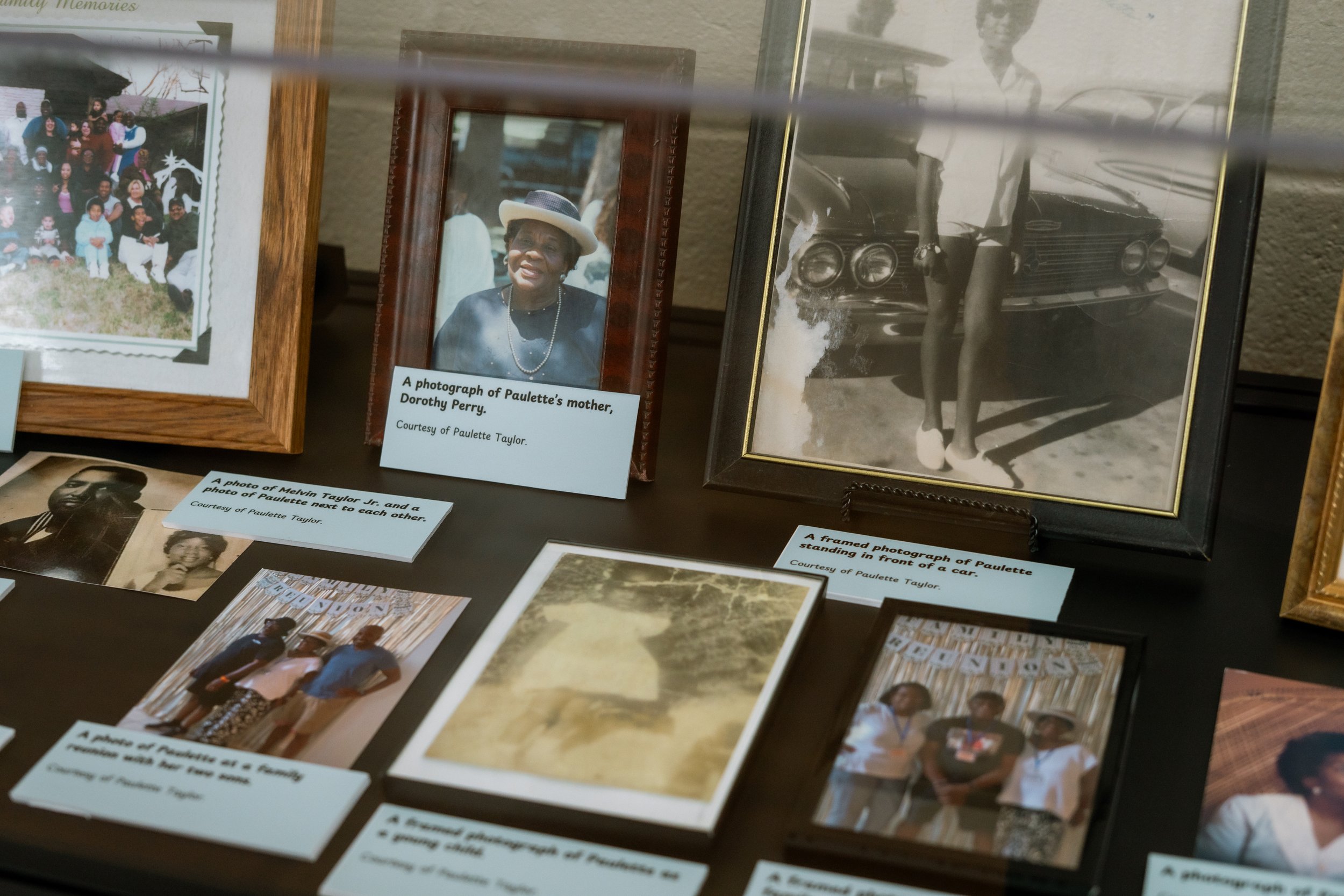 A collection of framed photographs and printed captions displayed on a table or wall, including a color portrait of an elderly woman wearing a hat, black-and-white photos of people, and images of cars and group gatherings.