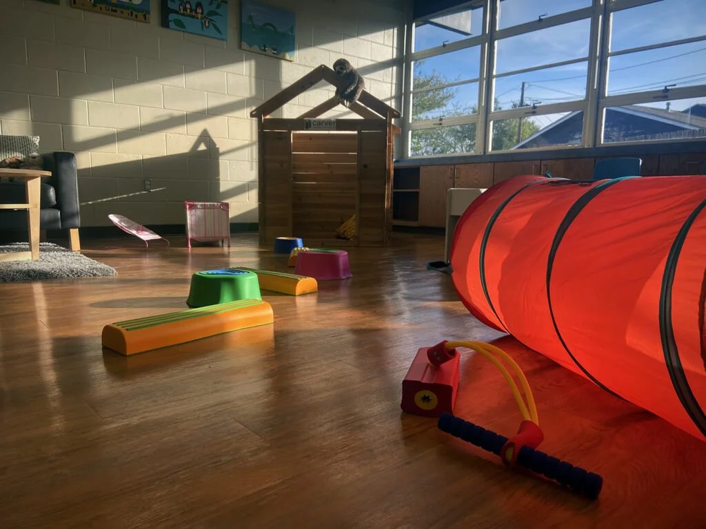 An indoor playroom with a large red tunnel, colorful stepping stones, a small batting cage, and various toys. Natural light streams through large windows, illuminating the wooden floor and wall art.