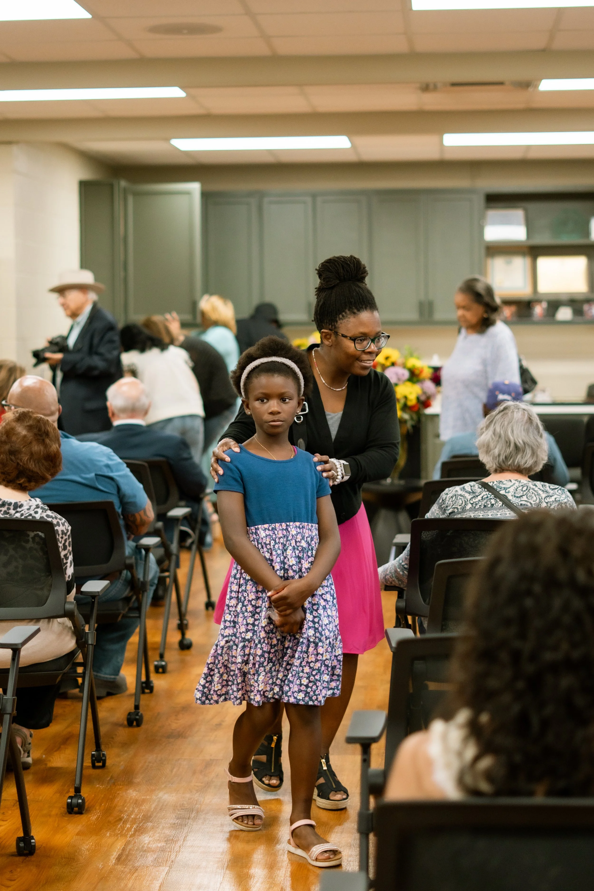 A woman and a young girl walk together in a room with many seated people. The woman has her hair in a large bun and wears glasses, a black top, and a pink skirt. The girl has curly hair with a headband, and is wearing a blue top and a floral skirt. The room has gray cabinets, a wooden floor, and a ceiling with rectangular lights. Other people are seen in the background, some standing and some seated, with a display of colorful flowers.