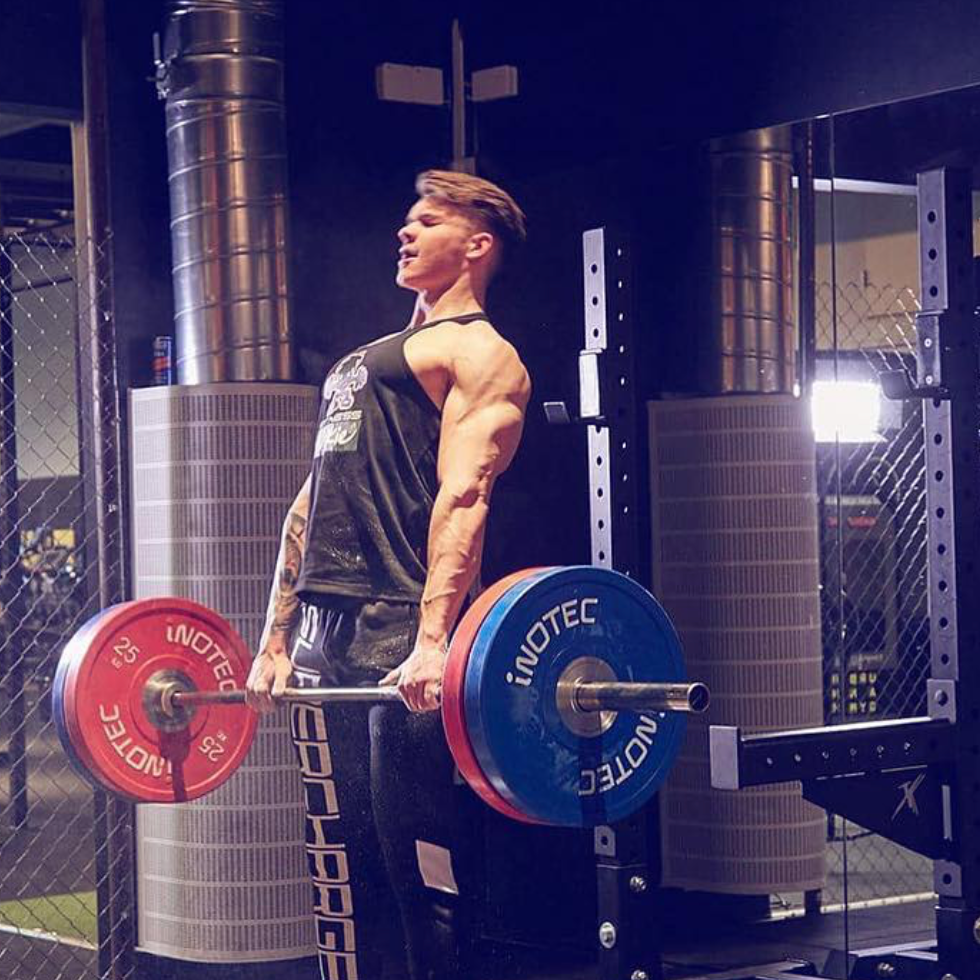 A woman with a muscular build lifting a barbell loaded with red and blue weight plates in a gym with industrial decor and a chain-link fence.