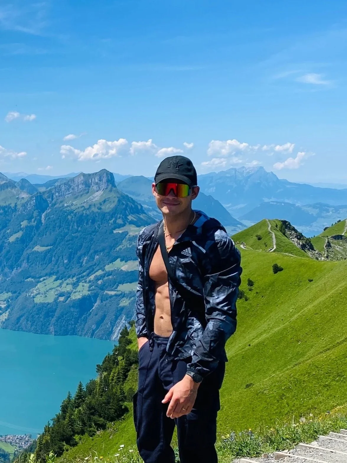 A man standing outdoors on a mountain trail with green hills and a lake in the background. He is wearing a black cap, sunglasses, a partially unzipped jacket showing his bare chest and abs, and black pants.