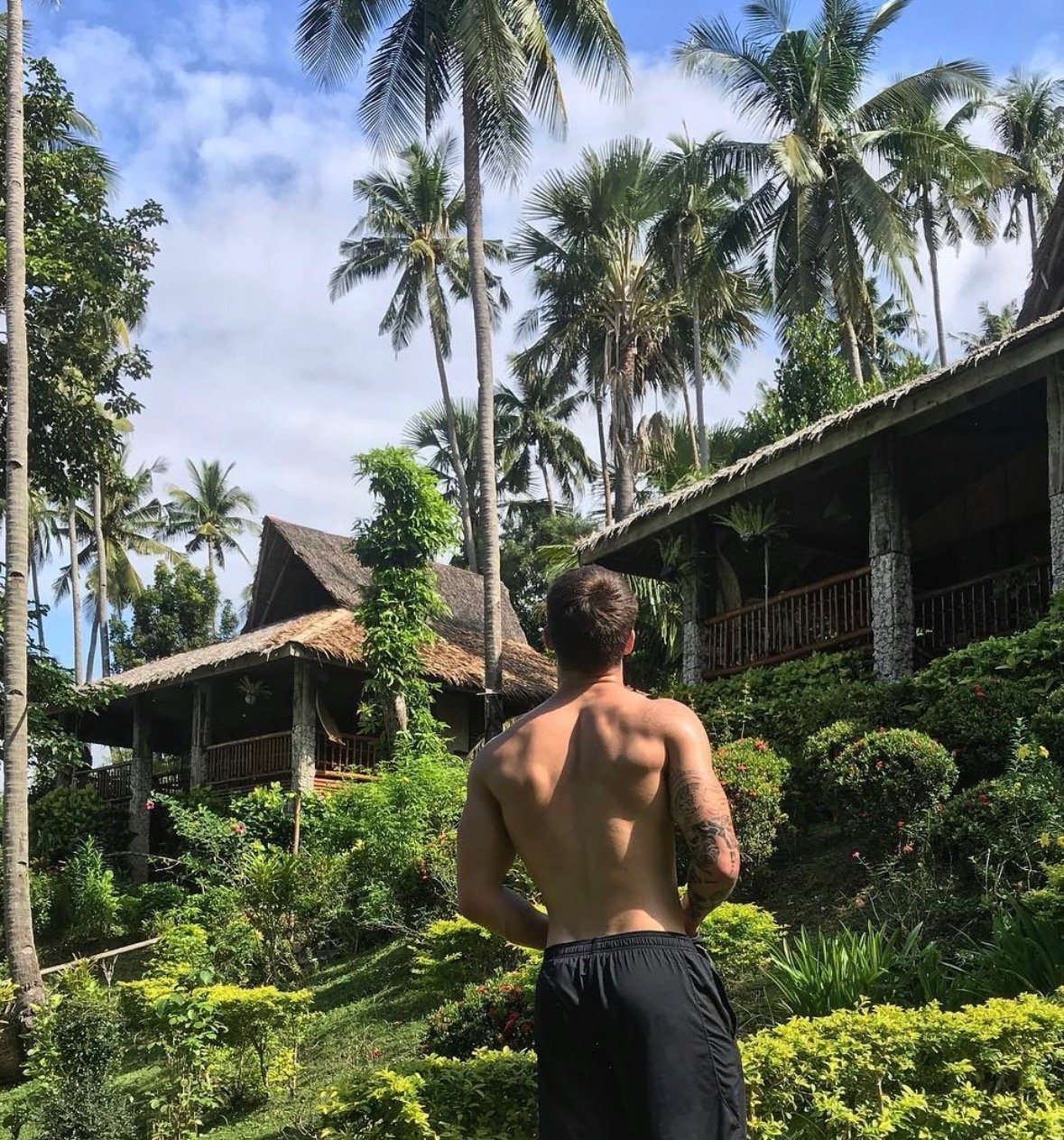 A shirtless man with tattoos standing in a tropical garden with palm trees and wooden huts.