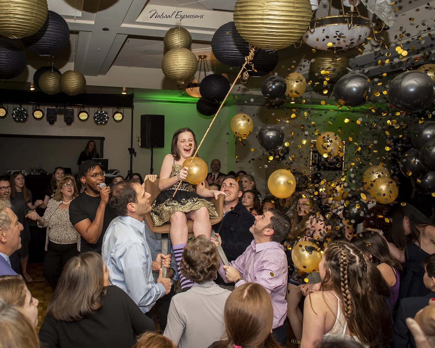 A crowd of people celebrating at a party with balloons and confetti, including a girl in a black and gold dress sitting on a chair held aloft by adults, smiling and holding a gold balloon, with decorations and lighting in the background.
