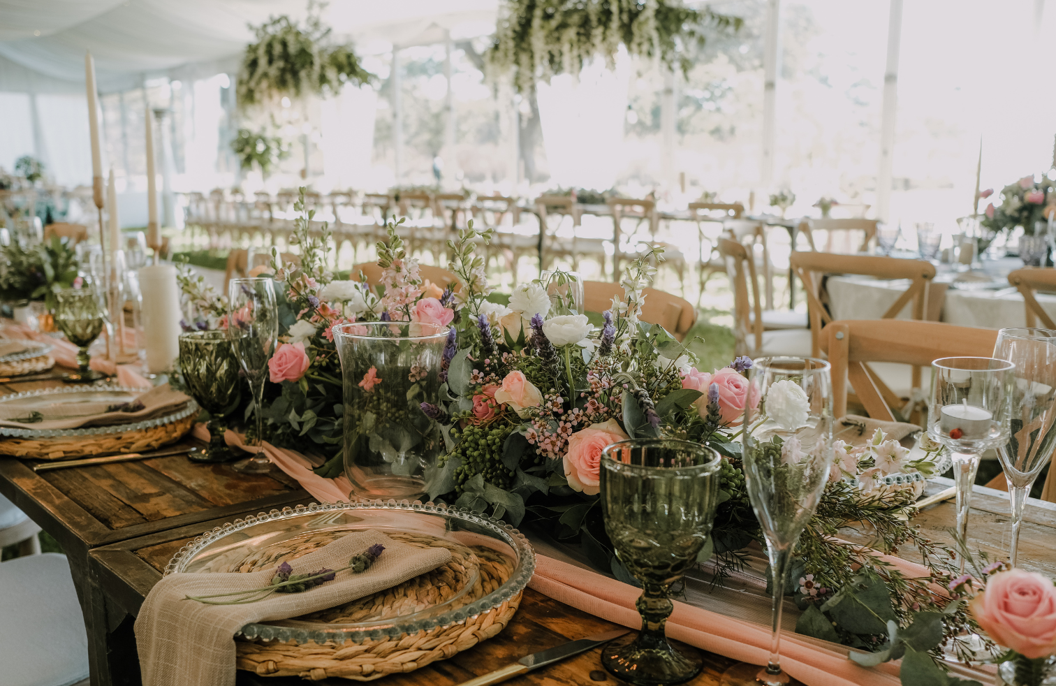 Elegant table decorated with a floral centerpiece of pink, white, and purple flowers, glassware, candles, and place settings in a bright outdoor tent.