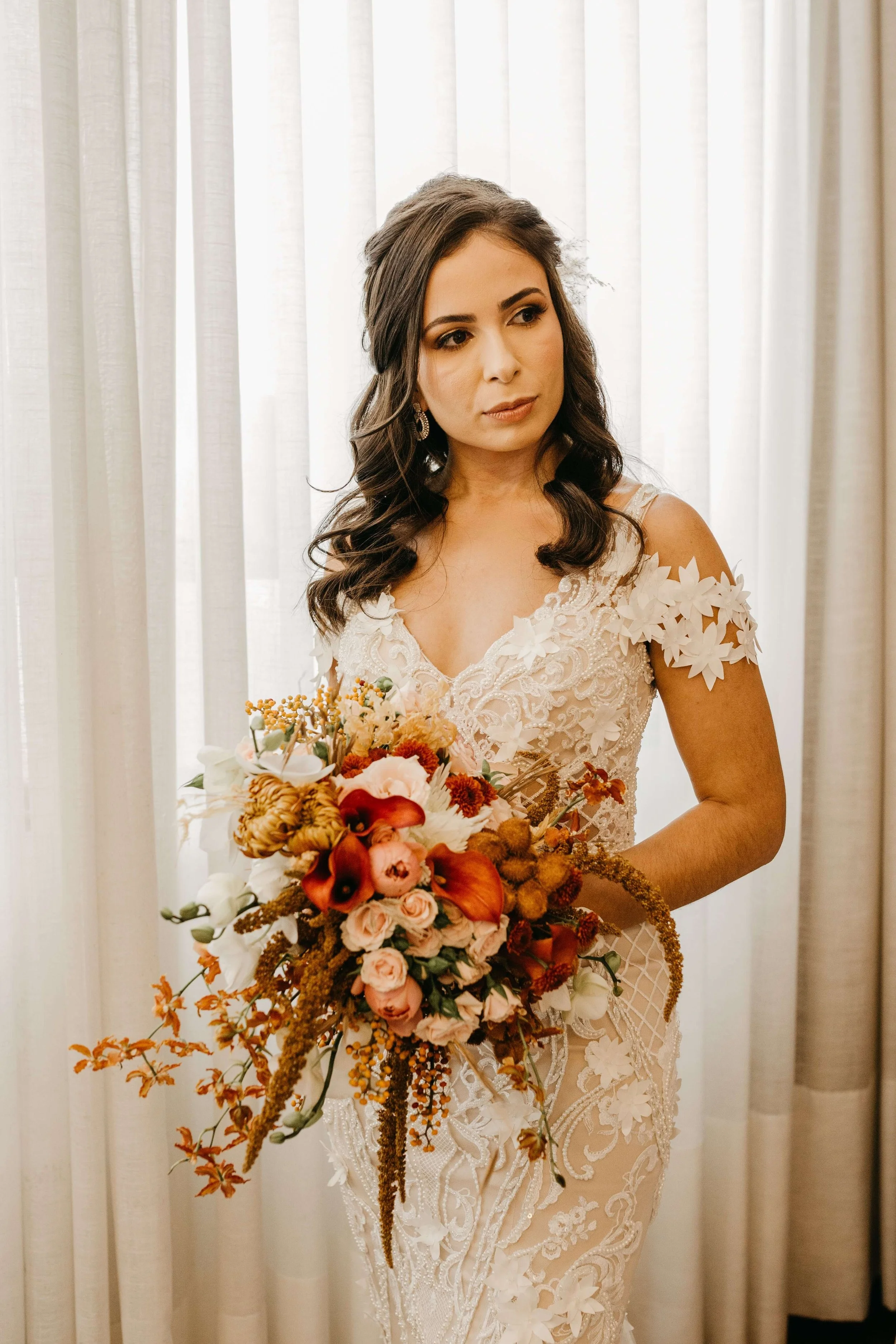 A woman in an elegant lace wedding dress holding a colorful bouquet of flowers, standing in front of a white curtain.