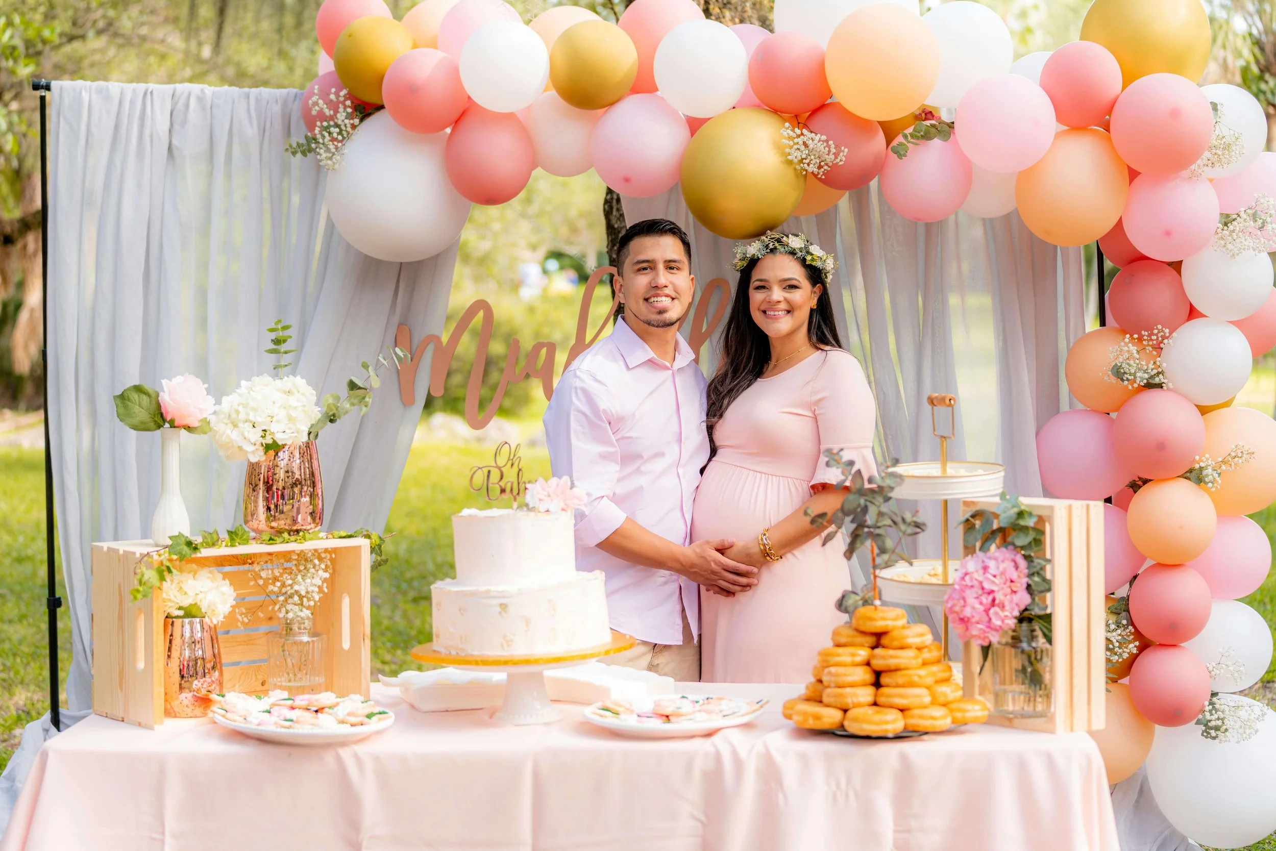 A pregnant woman and a man at a celebration table outdoors, surrounded by pink, white, and gold balloons, flowers, and desserts, during daytime.