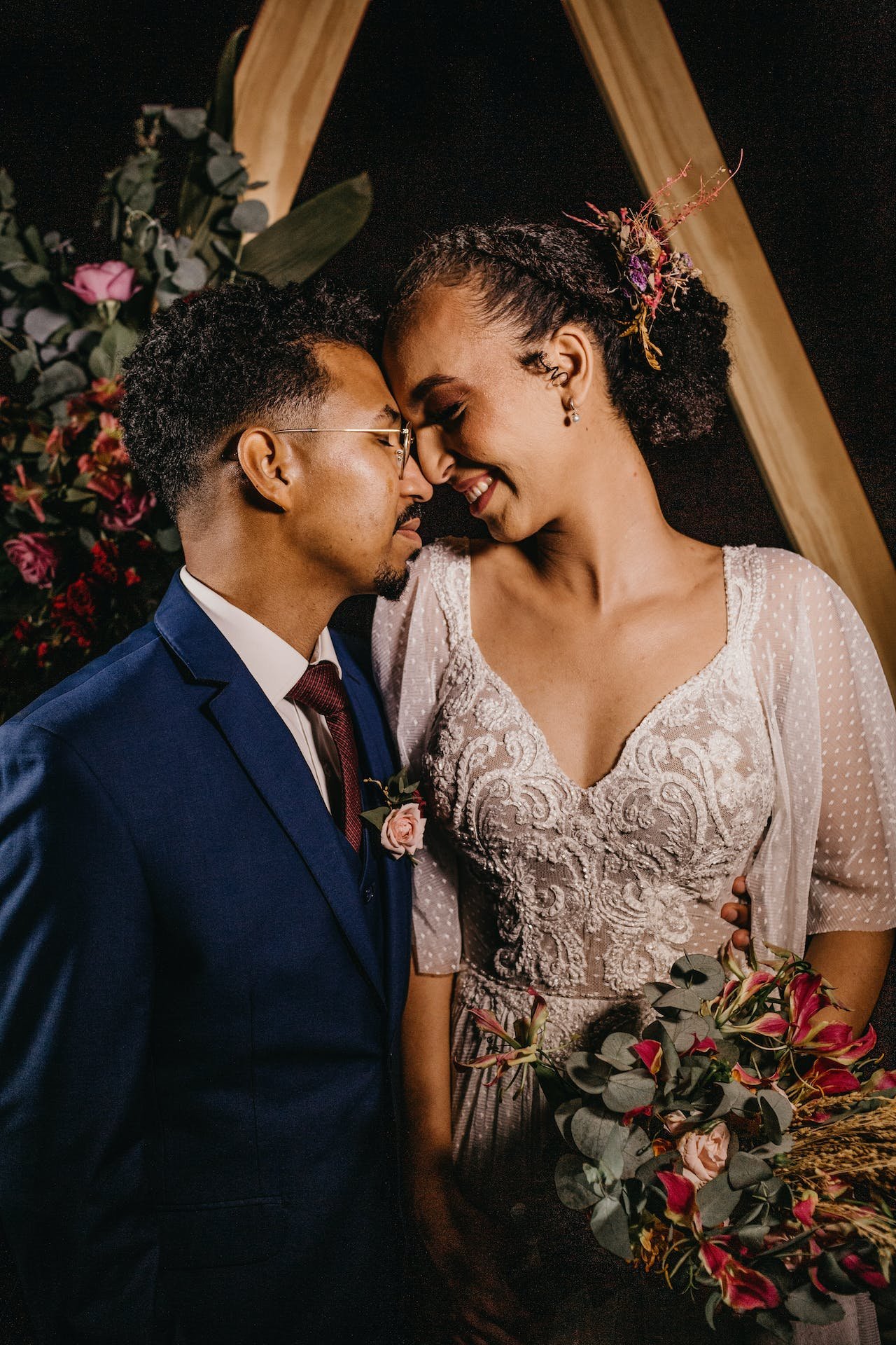 A couple touching foreheads at a wedding, with the man in a suit and the woman in a lace dress holding a bouquet of flowers.