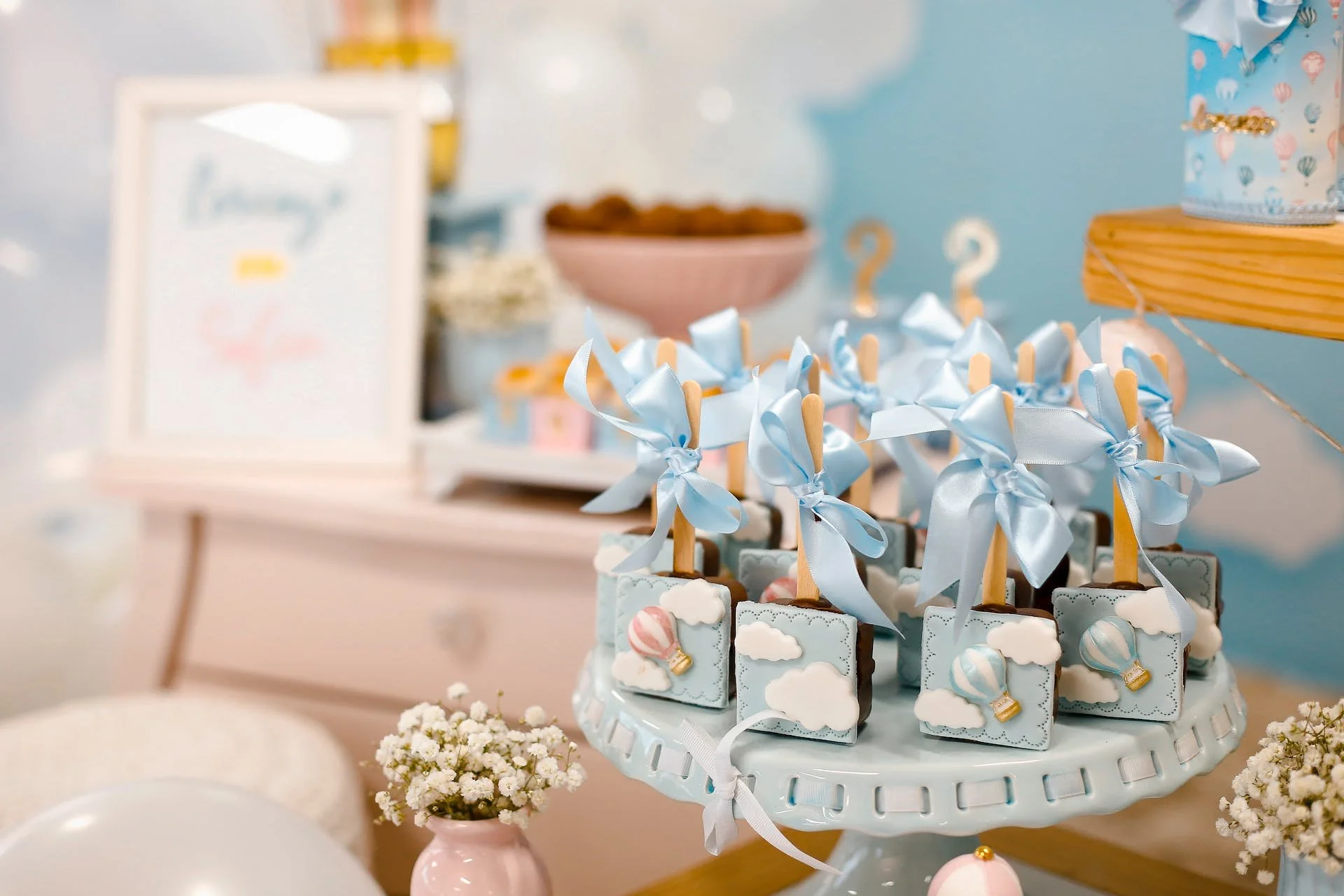 Baby shower party table with decorated cookies on a white cake stand, small white flowers in pink vases, and pastel decorations in the background.