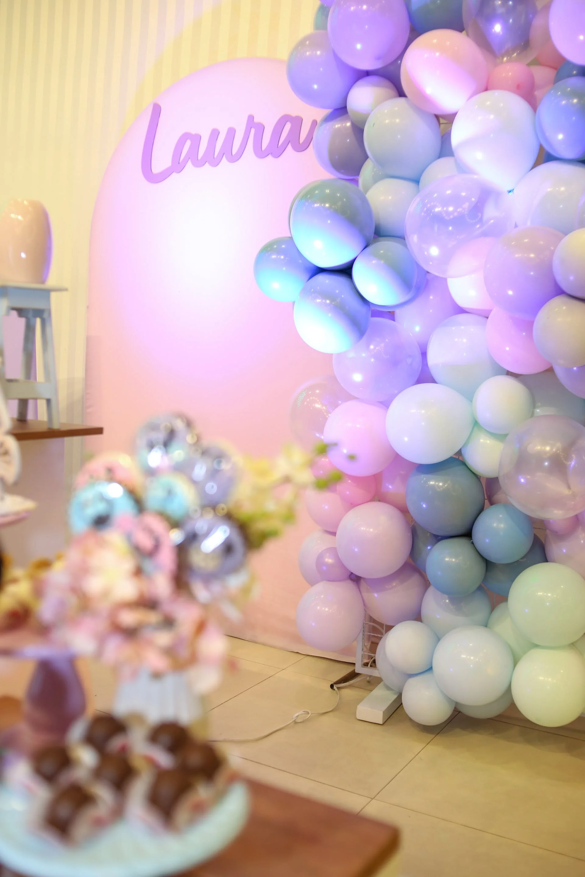 Decorative birthday setup featuring a backdrop with the name 'Laura' and a cluster of pastel-colored balloons in shades of purple, pink, and blue.