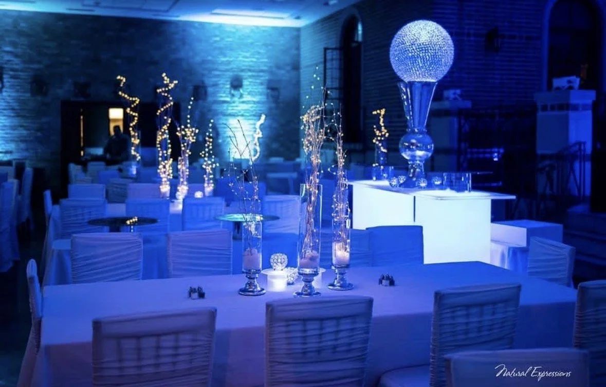 Elegant indoor event space decorated with tall glass vases and twinkling lights, illuminated with blue lighting, featuring a large decorative light fixture resembling a globe.