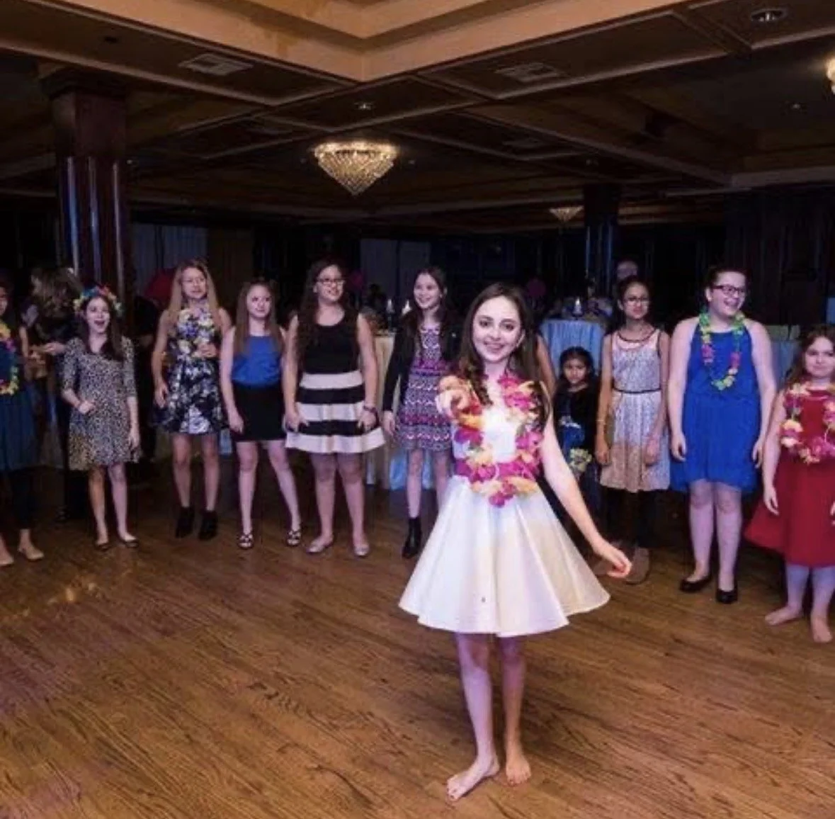 A group of girls dressed in party attire at a celebration, with one girl in the foreground wearing a white dress and a pink floral lei, smiling at the camera in a decorated indoor venue.