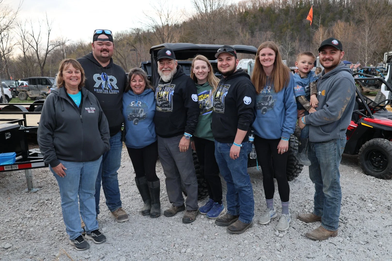 A group of nine people, including women and men of various ages, are standing outdoors on a gravel area in front of an off-road vehicle, with trees and a cloudy sky in the background. They are smiling and wearing casual or outdoor clothing, some with jackets and hoodies, and appear to be at a recreational or community event.