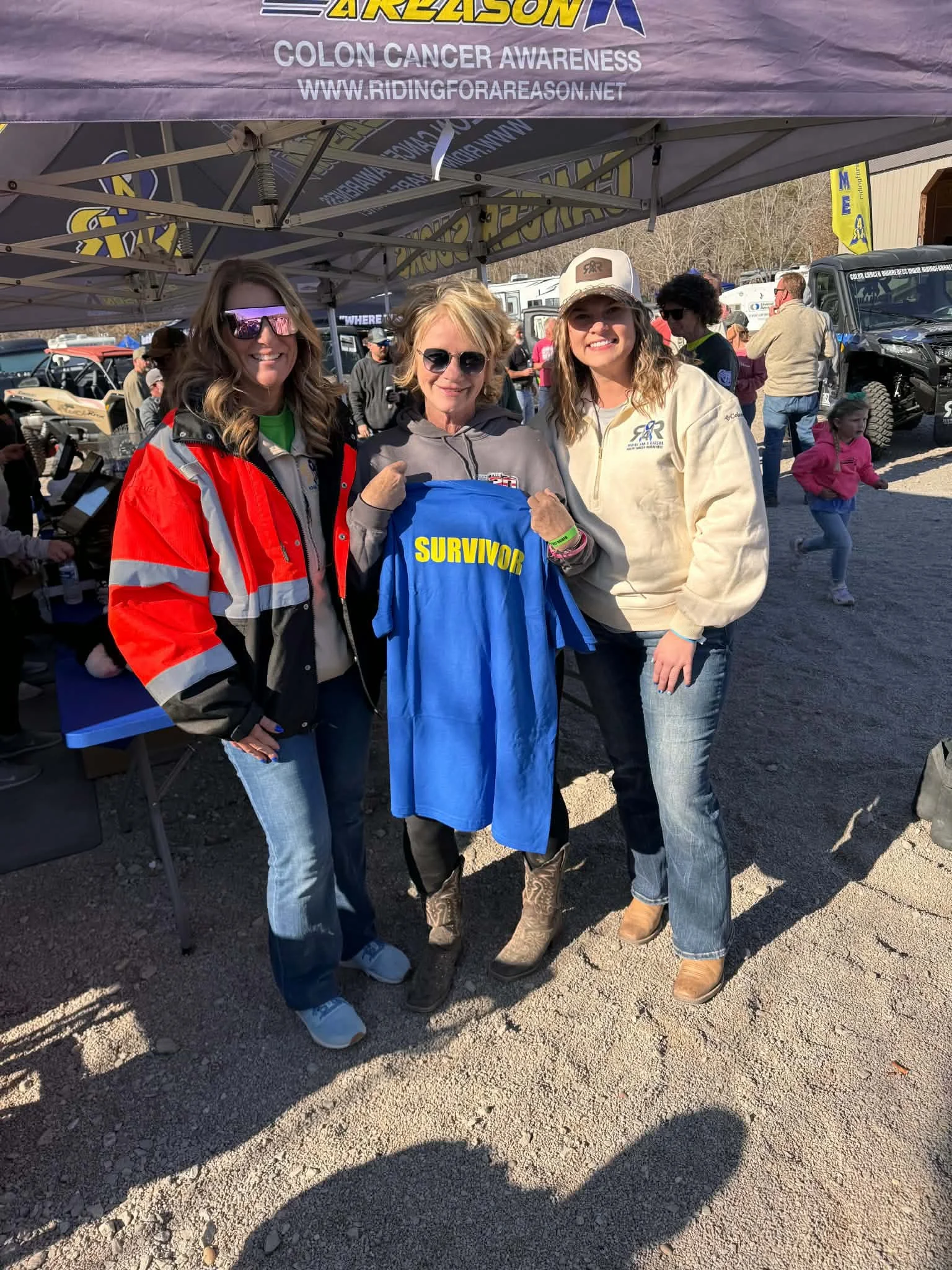 Three women standing together outdoors at a charity event for colon cancer awareness. They are smiling and holding a blue shirt with 'SURVIVOR' written on it. There are tents, vehicles, and other people in the background.