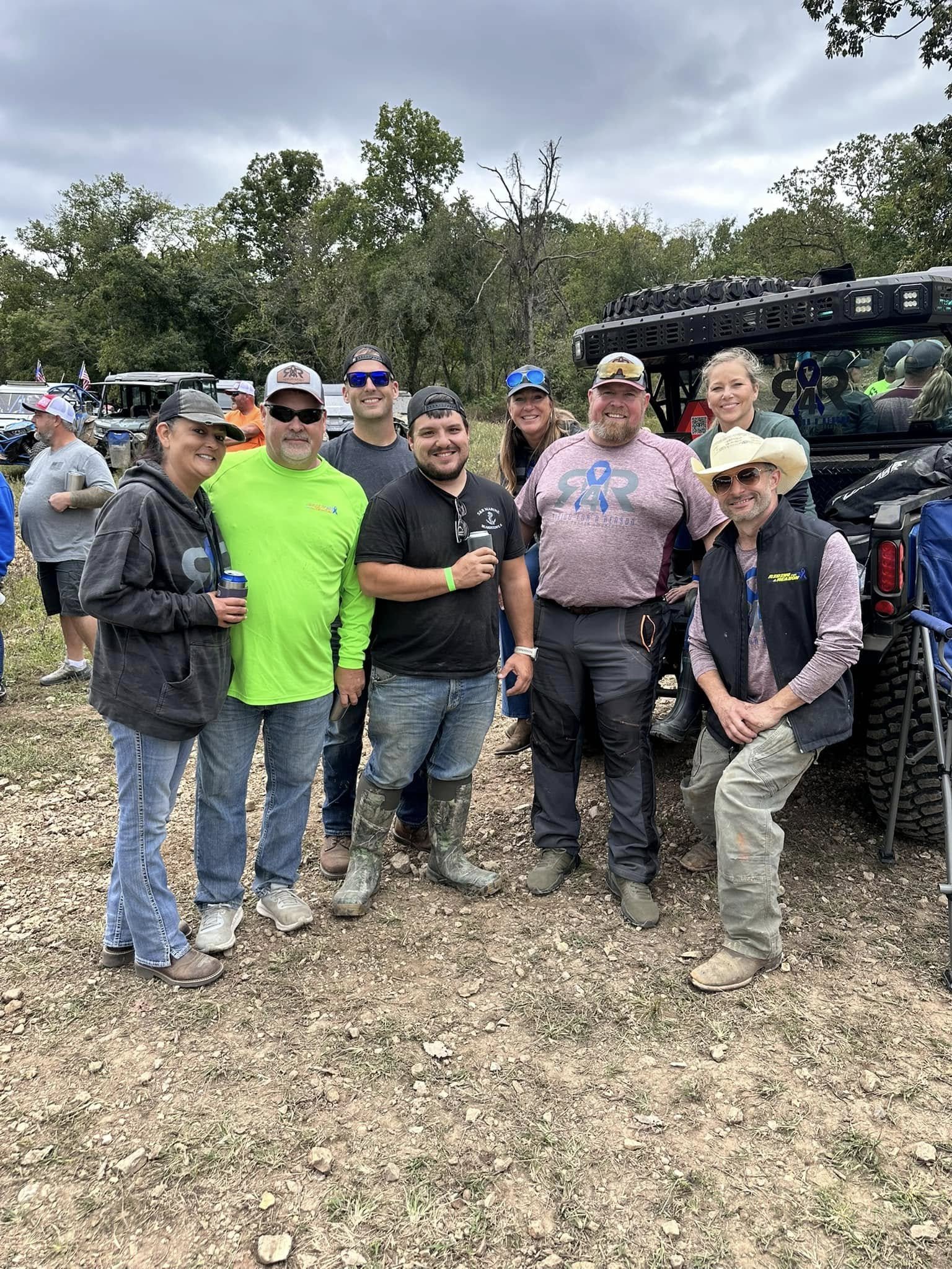 Group of smiling people outdoors in front of a large off-road vehicle with gear, trees, and a partly cloudy sky in the background at an outdoor event or gathering.