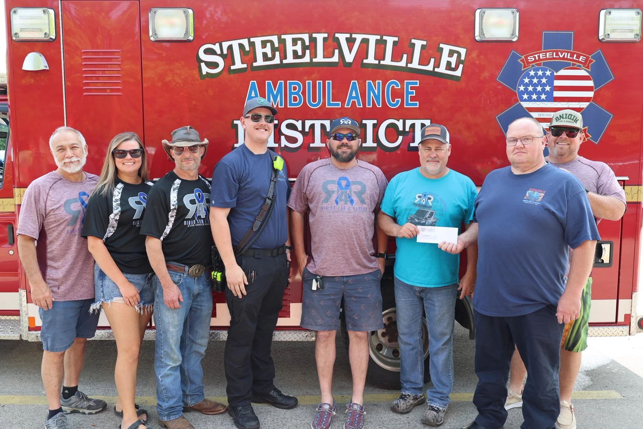 A group of nine people, including men and women, standing in front of a red Steelville Ambulance District vehicle. They are smiling and dressed casually, with some wearing sunglasses, hats, and t-shirts representing the ambulance service.