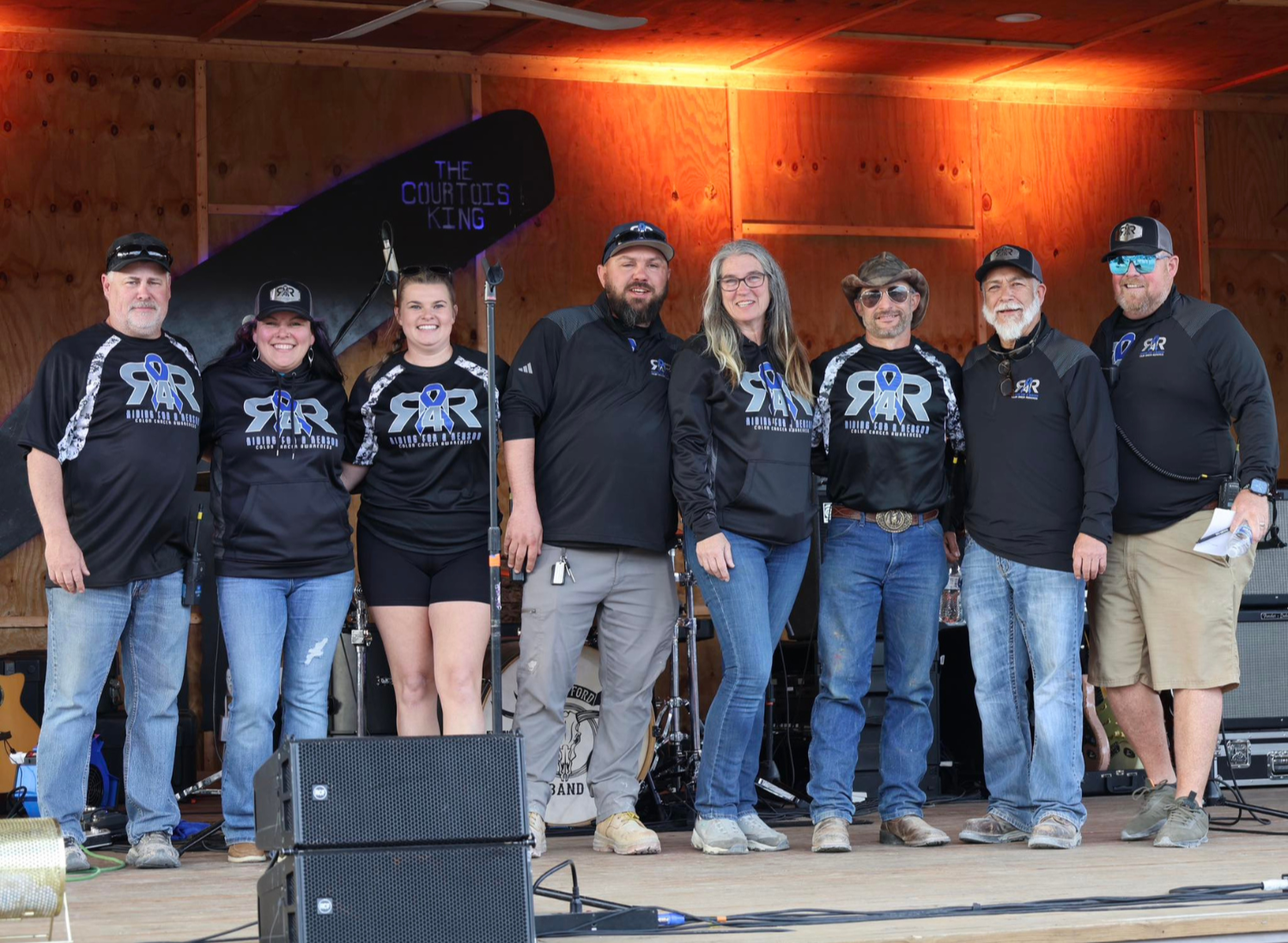 A group of eight people standing on a stage in front of a black bass guitar with the words "The Curious King" written on it, all wearing black shirts with a logo that says "SR" and a blue ribbon, smiling for a photo.
