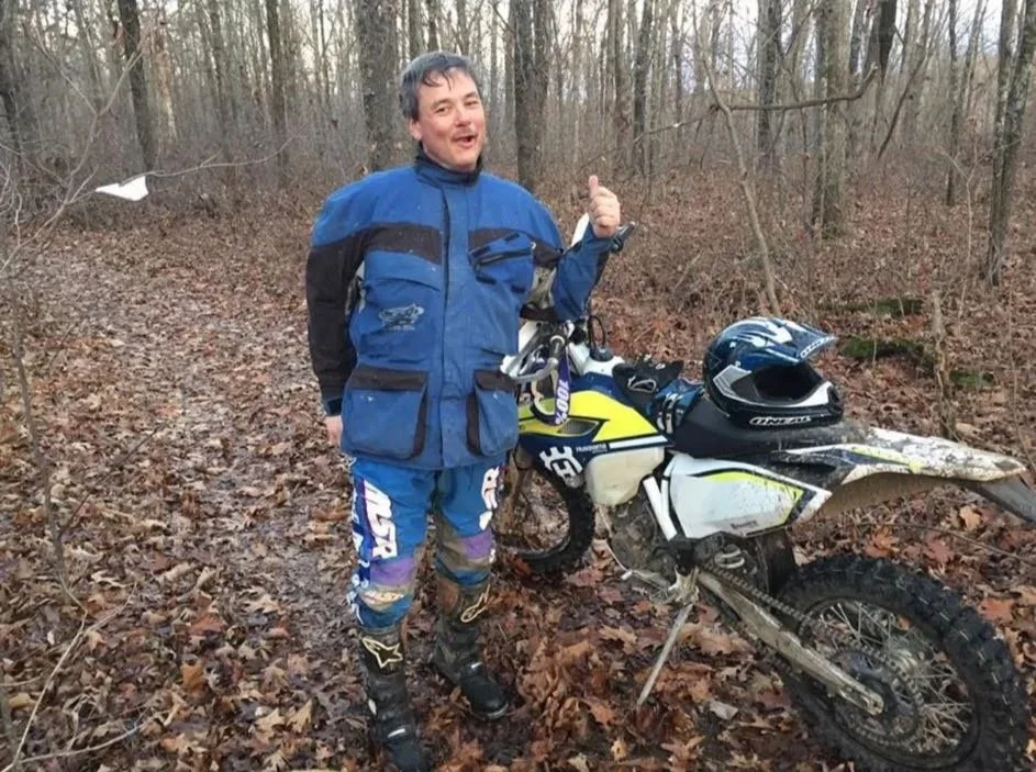 A man in motocross gear standing next to a dirt bike in a wooded area, giving a thumbs-up.