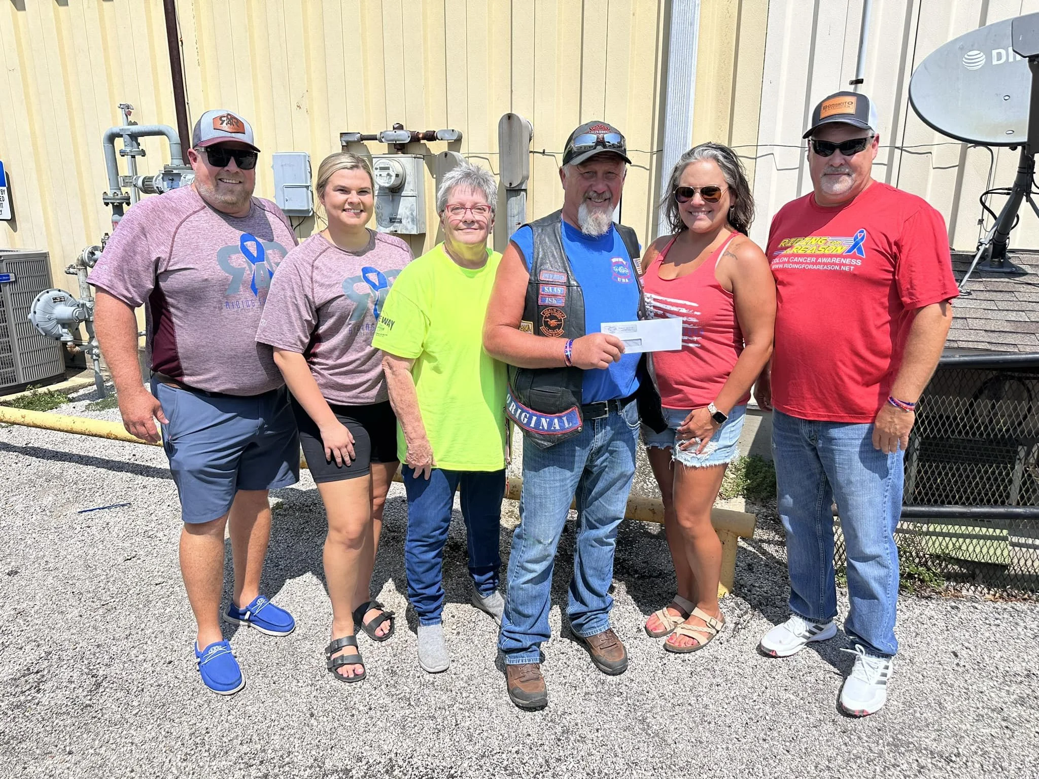 Group of six people standing outdoors in front of a yellow building, smiling at the camera. They are mostly wearing casual clothing, with some wearing sunglasses and hats. One person is holding an envelope or check. The background includes utility meters and pipes.