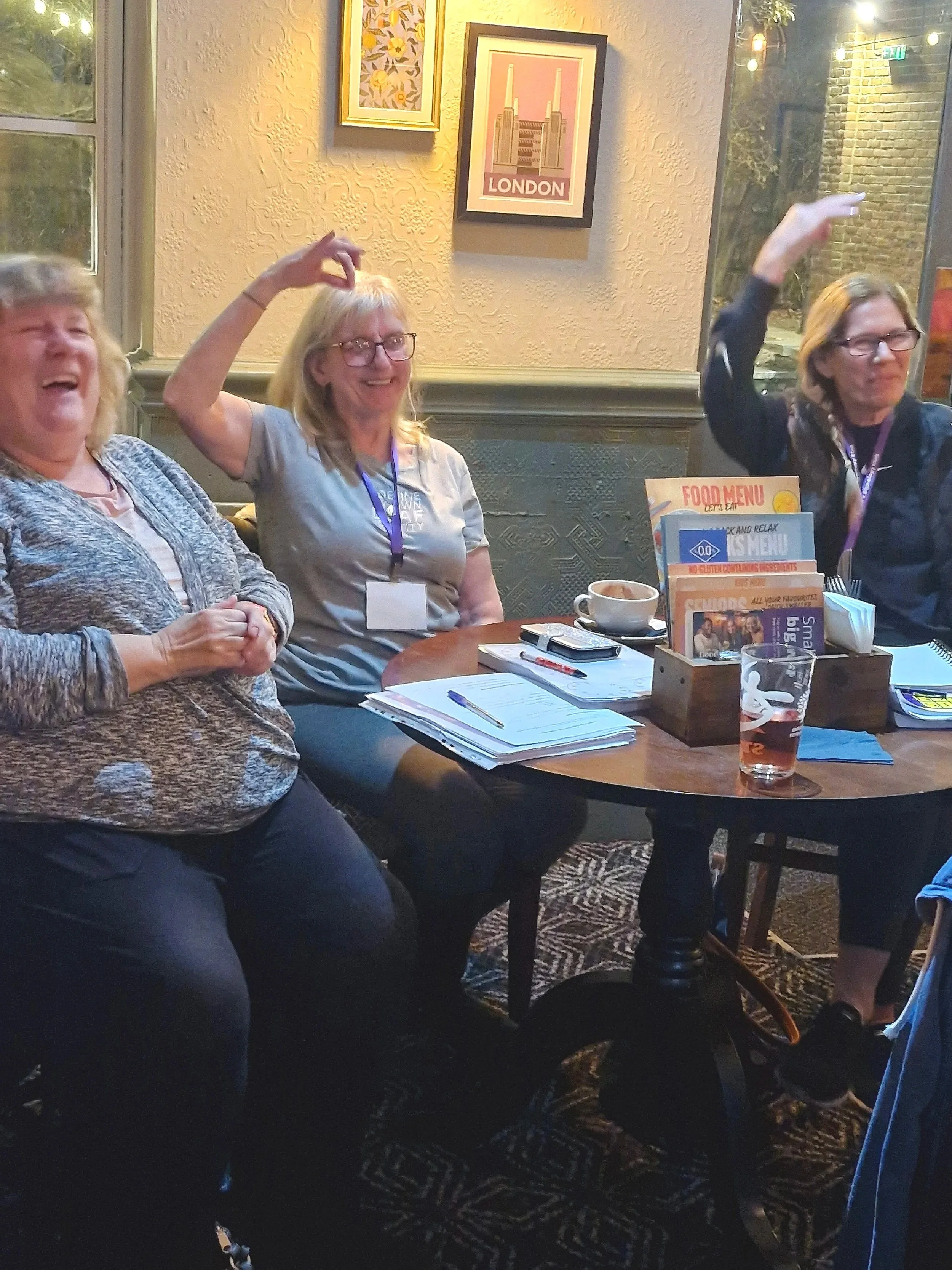 The image shows three female students practicing a multi-channel BSL sign. The lady of the left is laughing whilst the other two smile and practice.