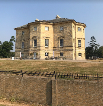 Image shows a picture of Danson House on a dry, sunny day. The sky is clear blue, the grass in the foreground is faded and slightly sun scorched.