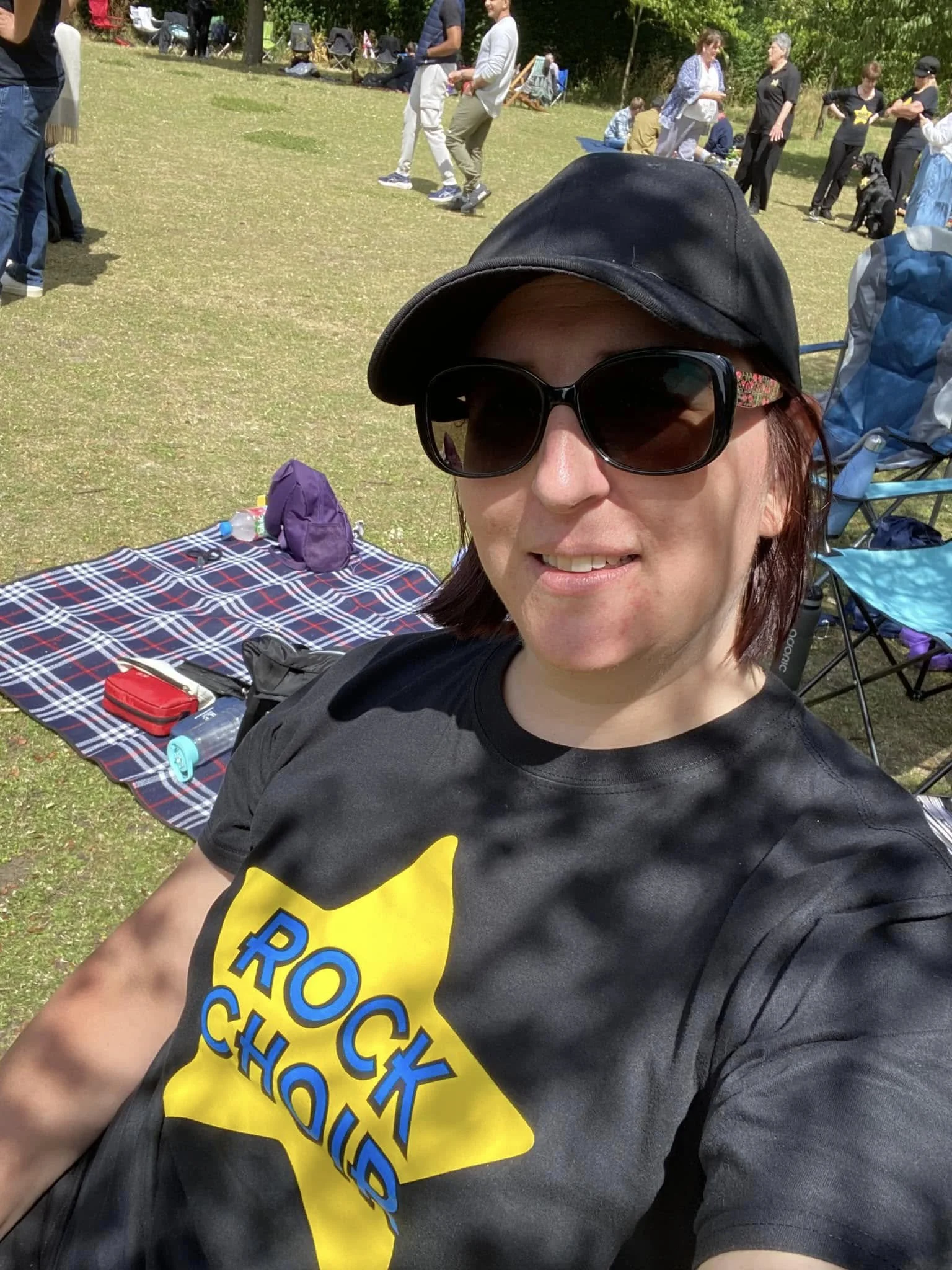 Smiling team member wearing sunglasses, a black t shirt with a yellow star with the words Rock Choir and a cap outdoors on a sunny day. She is sitting in front of a picnic blanket on a chair.