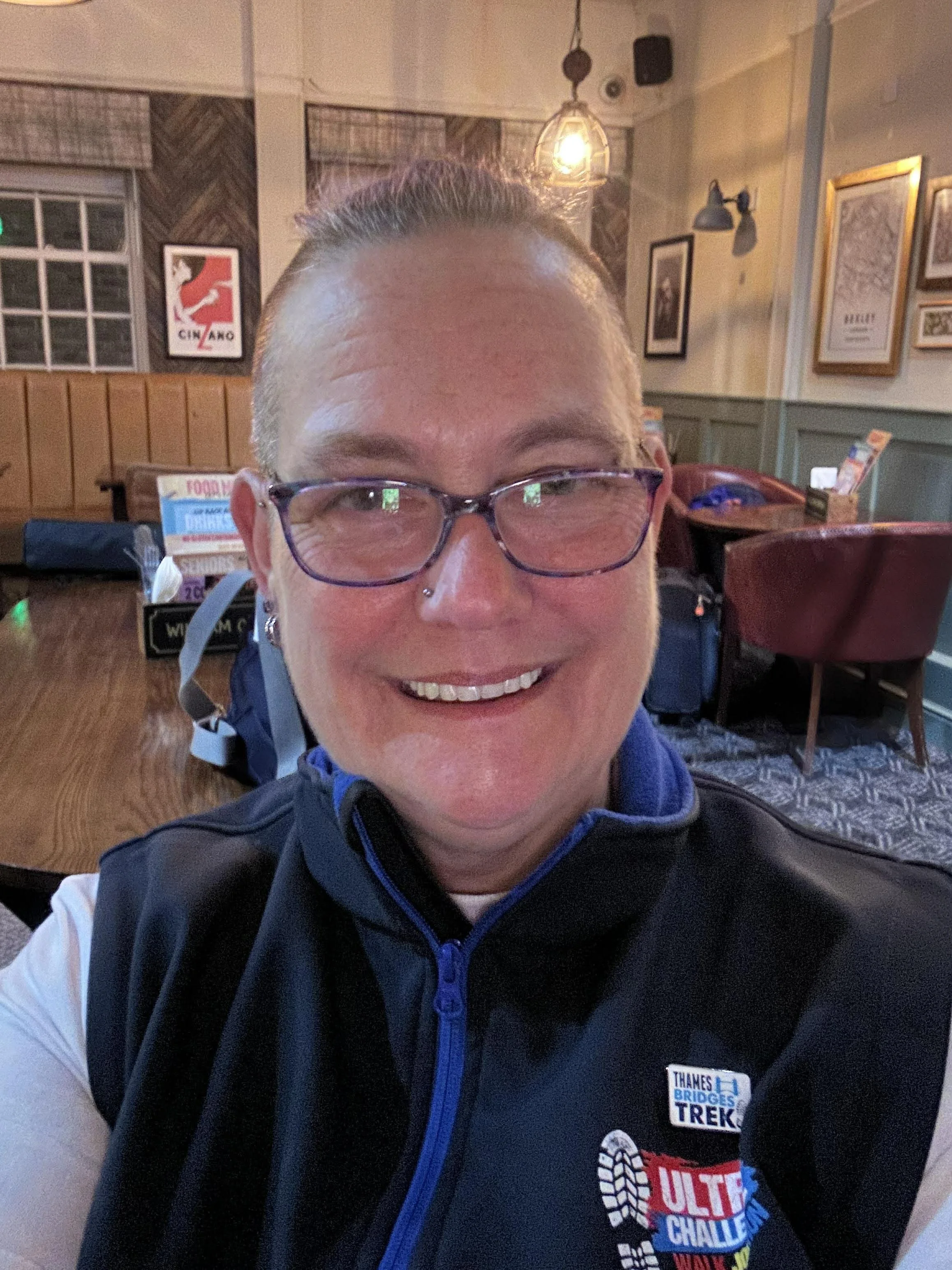 The image shows Smiling person wearing glasses and a dark sleeveless jacket, indoors in a cosy café with wooden furniture and framed artwork in the background.