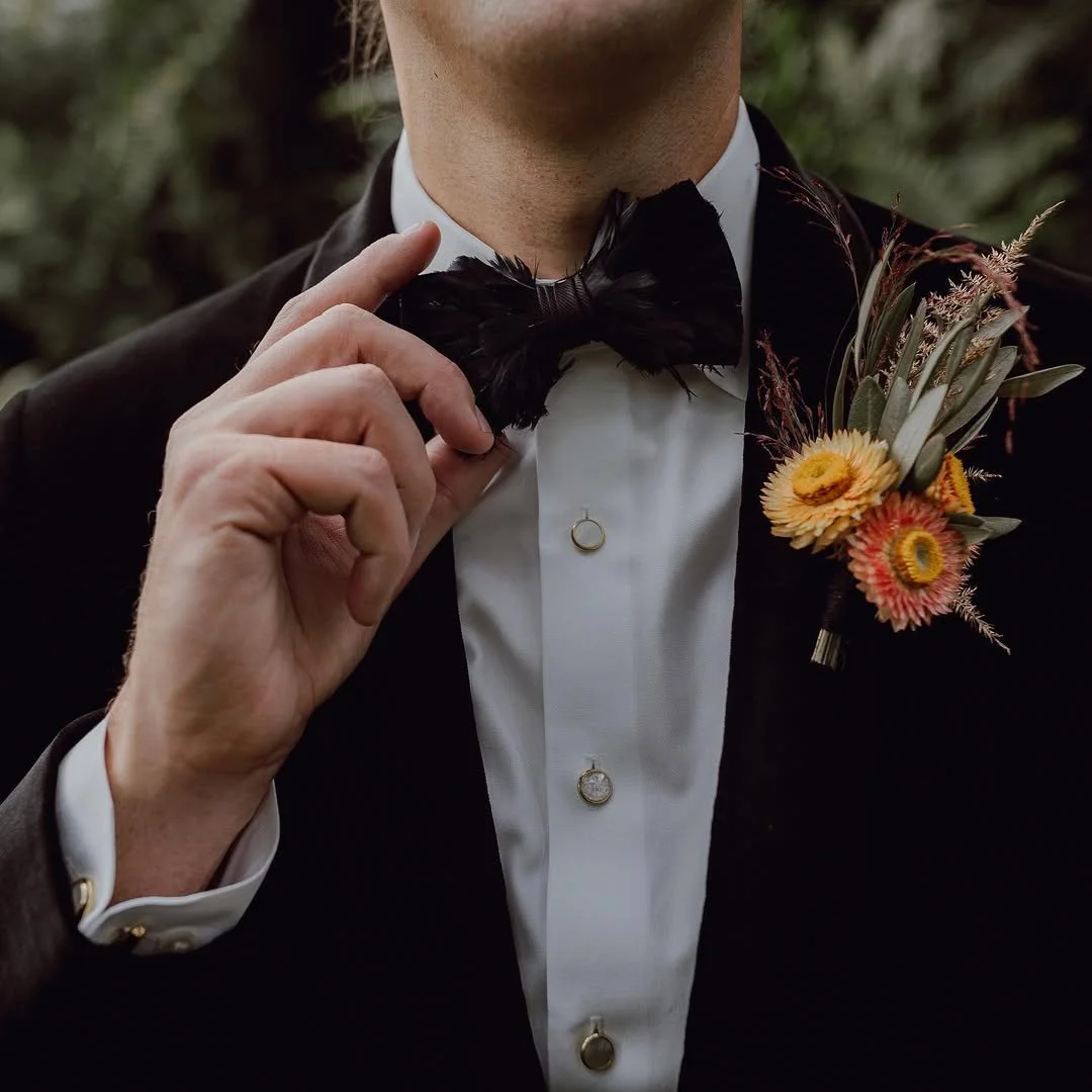 A man dressed in a tuxedo with a black bow tie, white shirt, and boutonniere with yellow and pink flowers. He is adjusting his bow tie with one hand.
