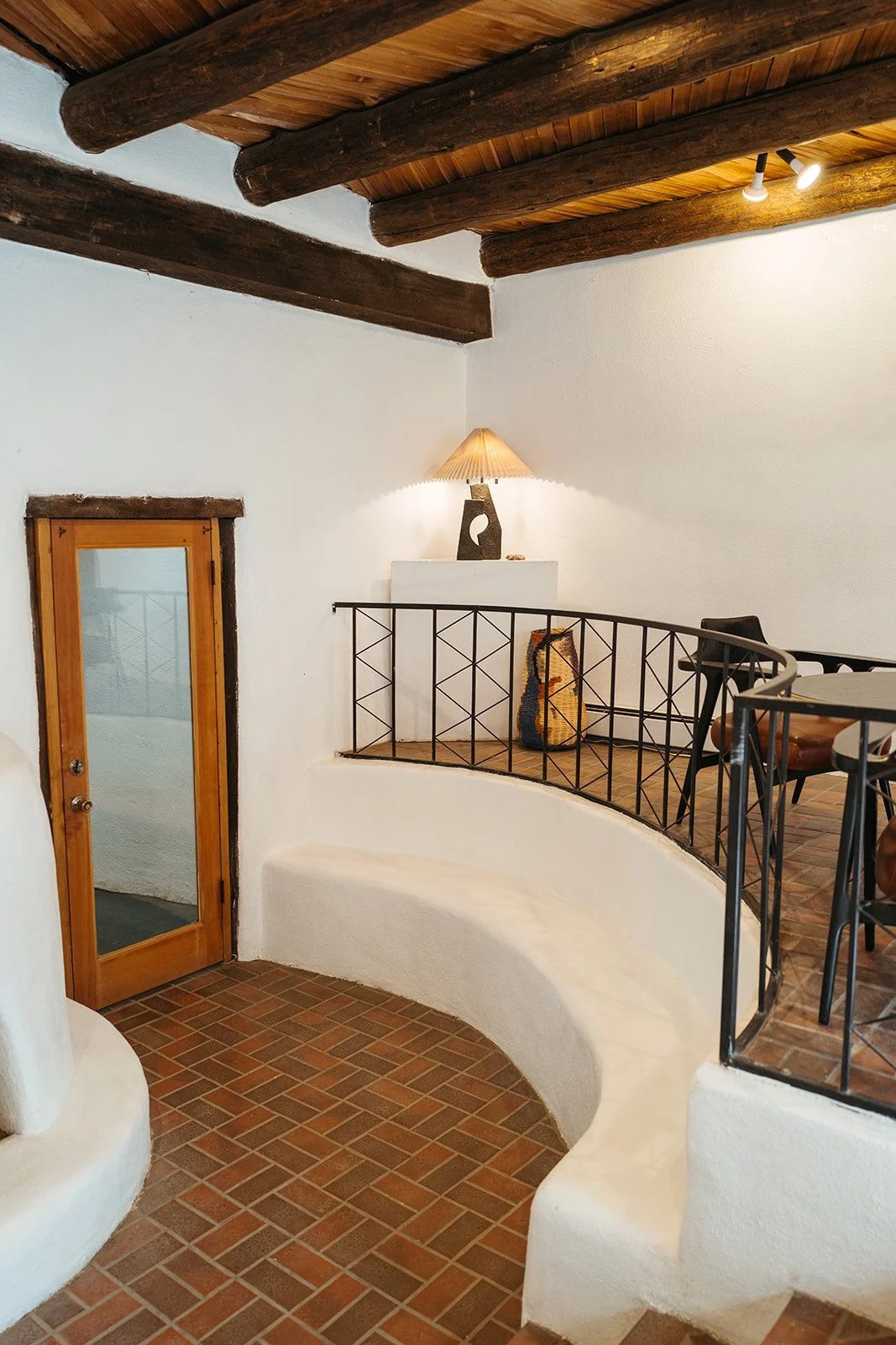 Interior of a room with white stucco walls, dark wooden beams on the ceiling, a wooden door with glass, a curved seating area, a small balcony with a black metal railing, a table with chairs, a table lamp on a white ledge, and a decorative woven bag.